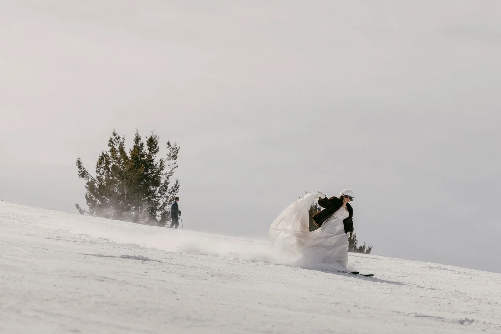  A person in a white wedding dress skis down a snowy slope, leaving a trail of snow, with trees and another skier visible in the background under a cloudy sky. 