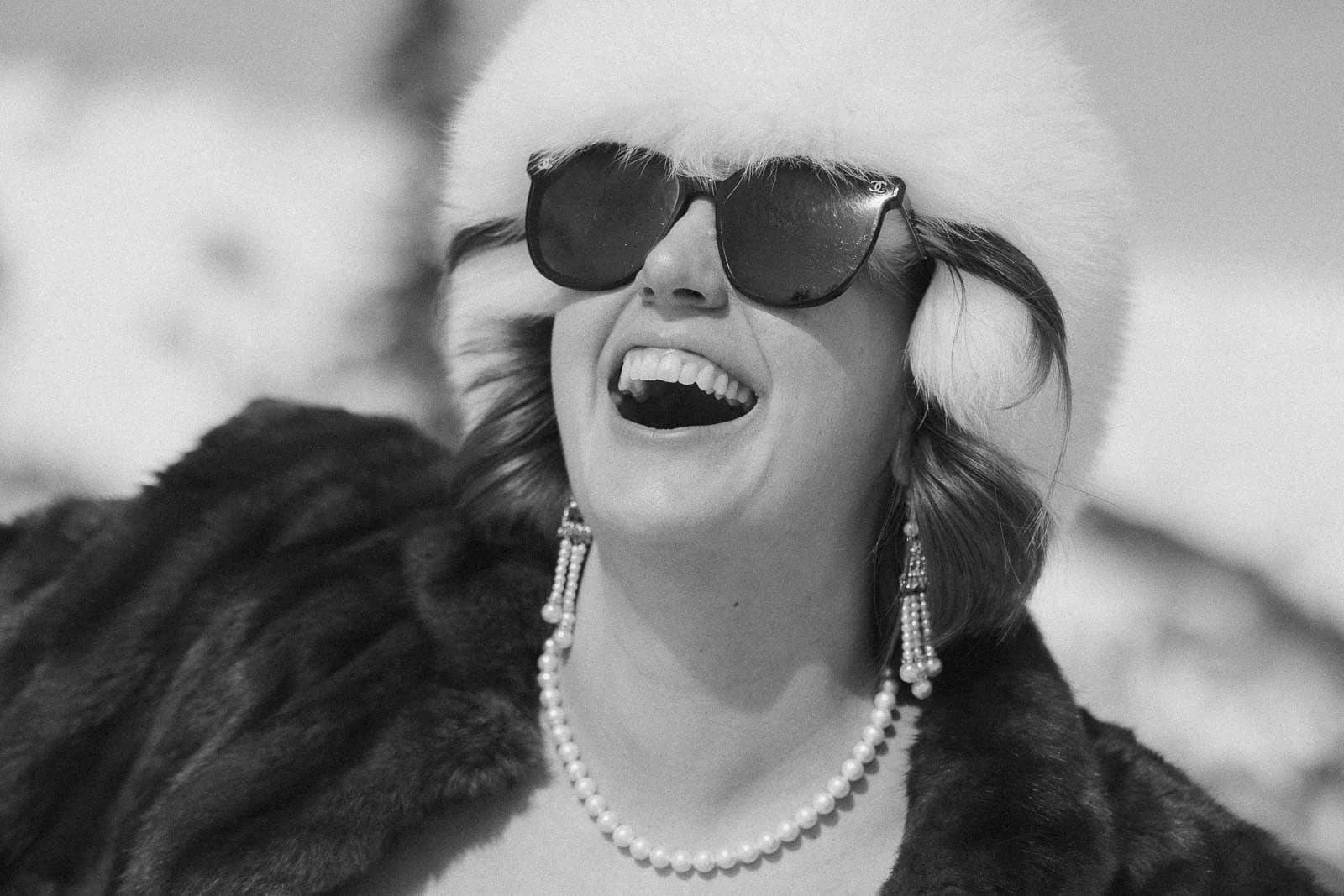  A woman wearing dark sunglasses, a fluffy white hat, dangling pearl earrings, and a pearl necklace laughs joyfully. She is dressed in a dark fur coat, and the background shows a snowy outdoor setting. The photo is black and white. 