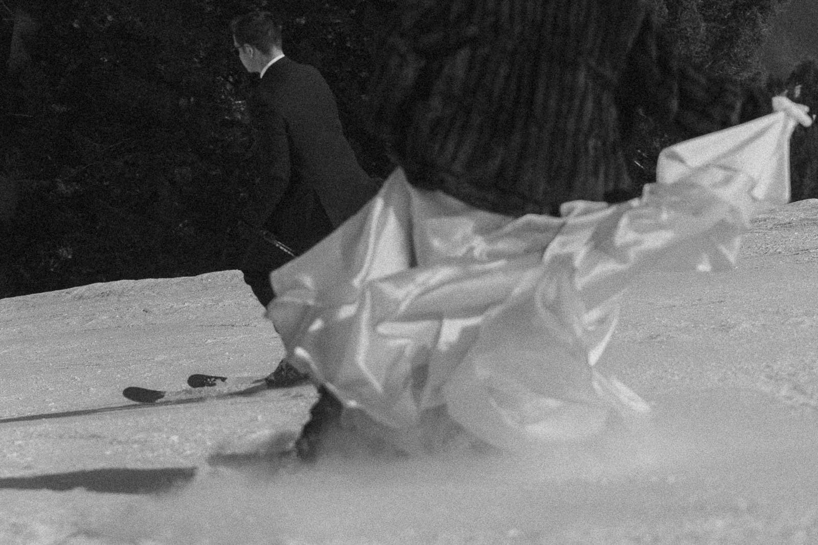  A black and white photo shows two people skiing downhill; one in a suit, the other in a long flowing dress, with the dress billowing as they move through the snow. 
