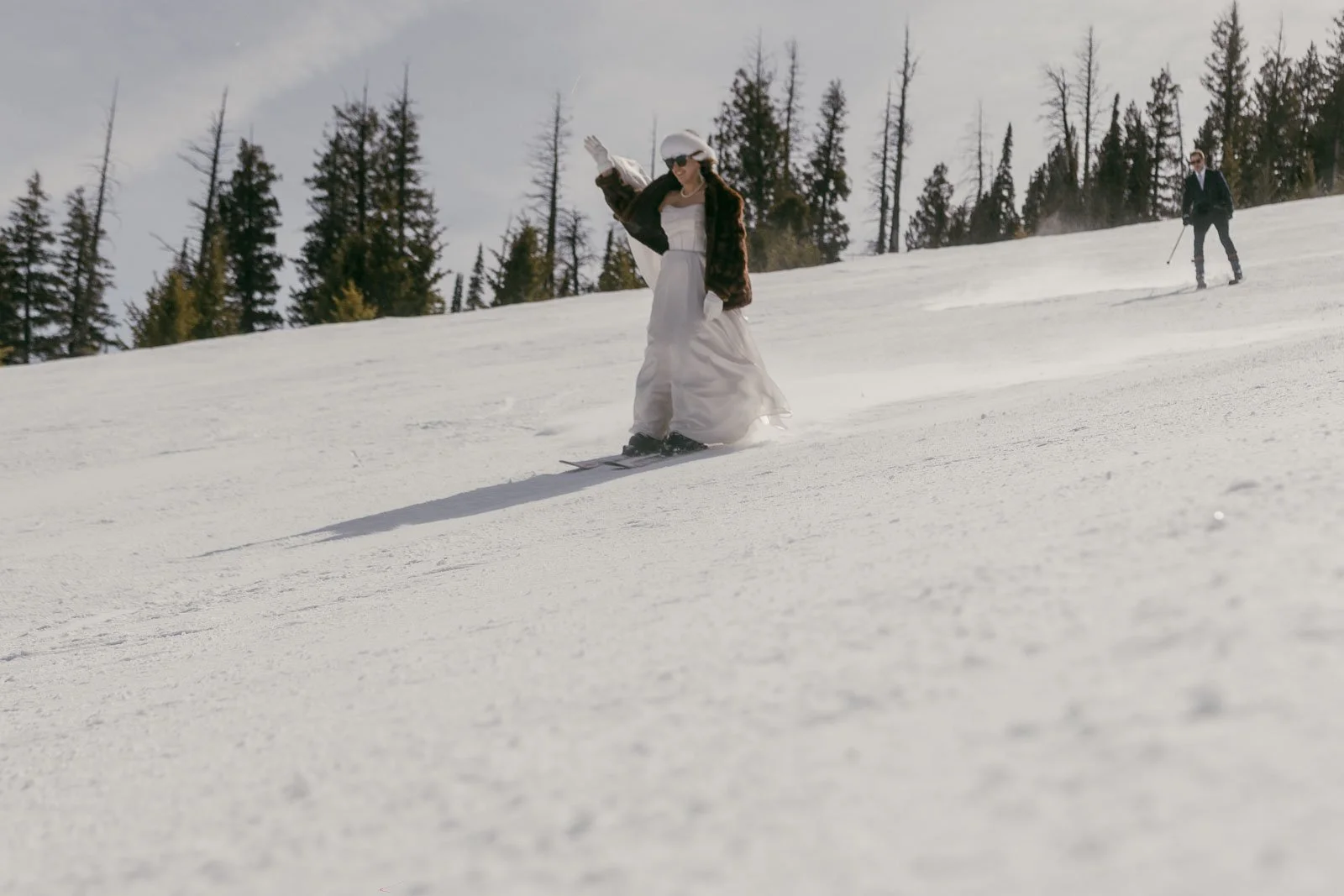  A person in a white wedding dress and veil skis down a snowy slope, with another skier in the background and pine trees along the horizon. 