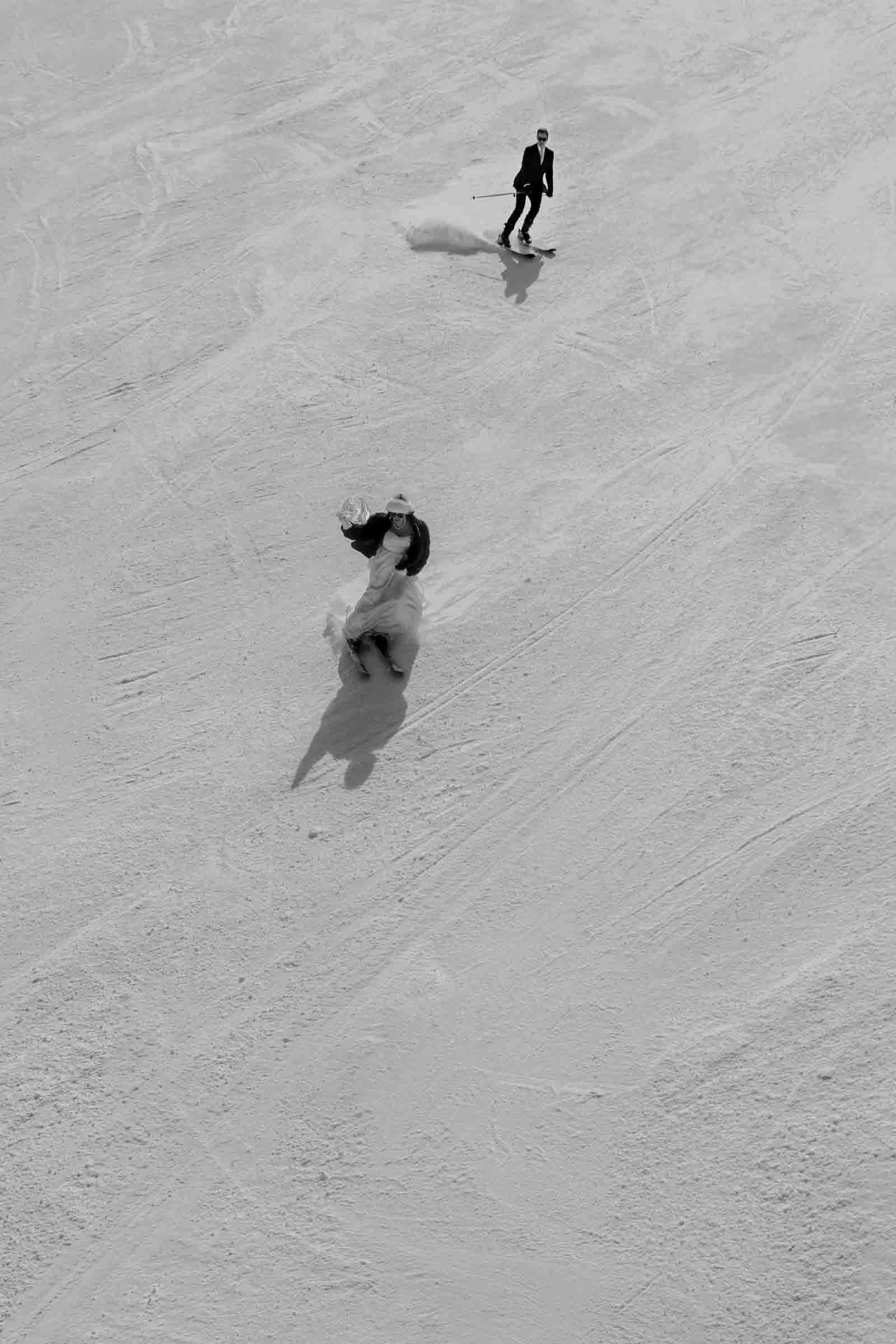 A bride in a flowing white dress and a groom in a suit ski downhill across a snowy slope, leaving tracks behind them in the snow. Their long shadows stretch across the landscape in this black-and-white aerial photograph.