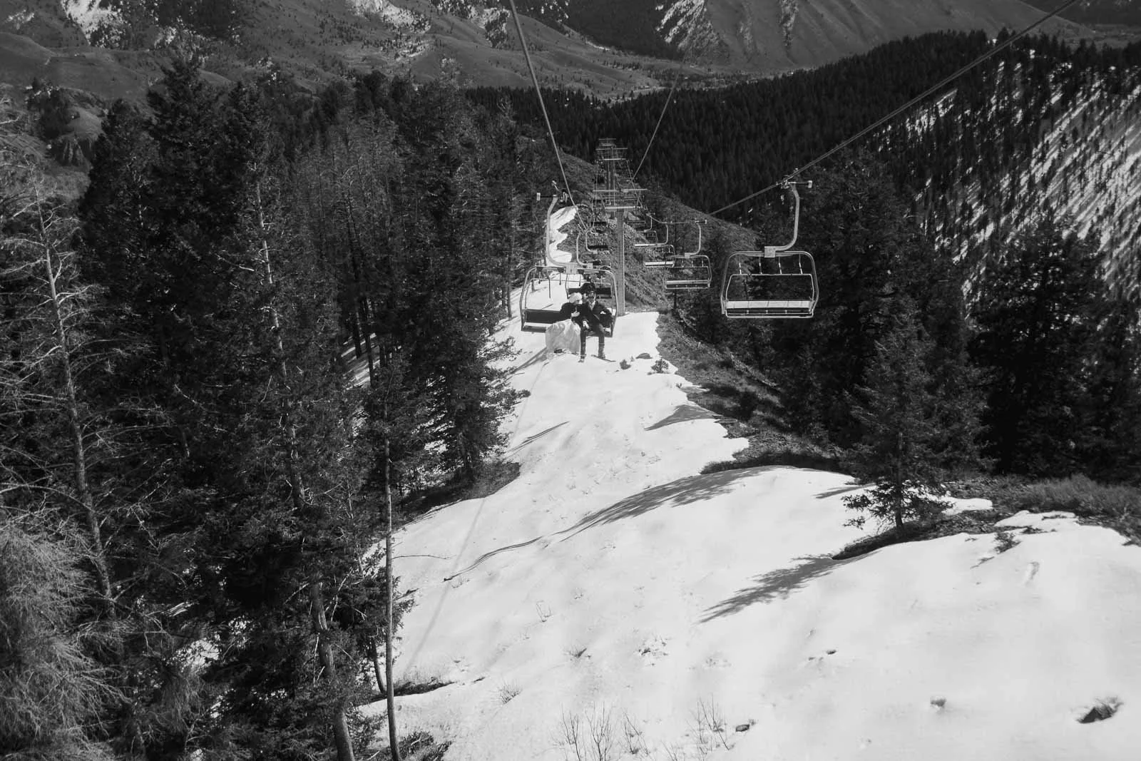  A ski lift carries people up a snowy mountain slope, surrounded by tall trees and rugged terrain, with mountains visible in the background. 