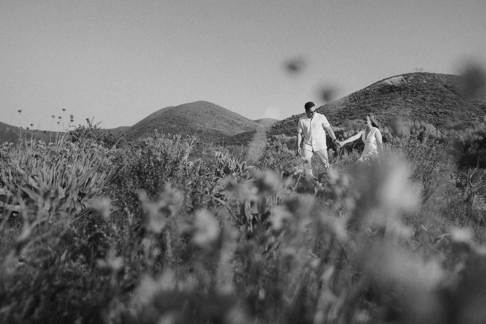  A couple holds hands while walking through a wildflower field, surrounded by hills under a clear sky. The image is in black and white, with blurred flowers in the foreground. 