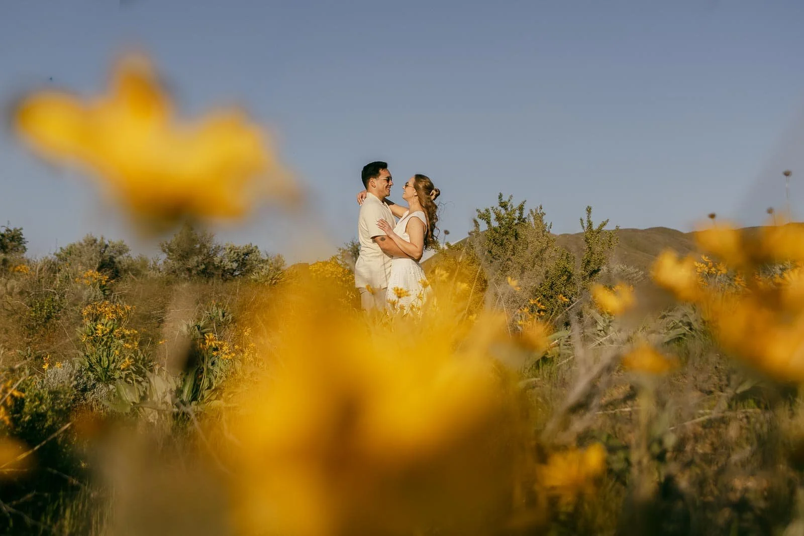  A couple stands embracing and smiling at each other in a field with large yellow flowers in the foreground, blurred for artistic effect, under a clear blue sky. 