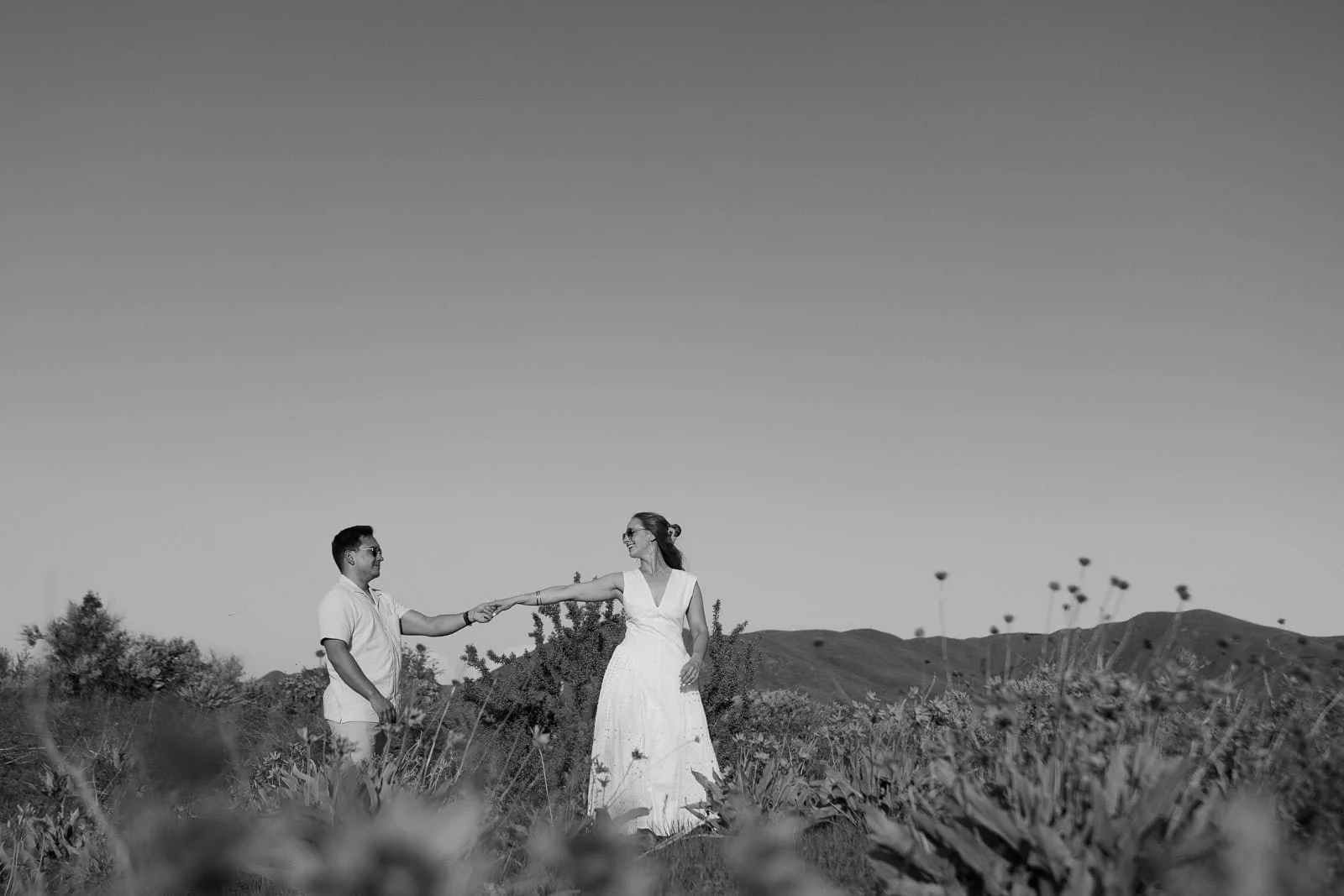  A couple stands in a field of wildflowers, holding hands and smiling at each other. The woman wears a white dress, and the man wears light clothing. Hills and a clear sky are visible in the background. The photo is in black and white. 