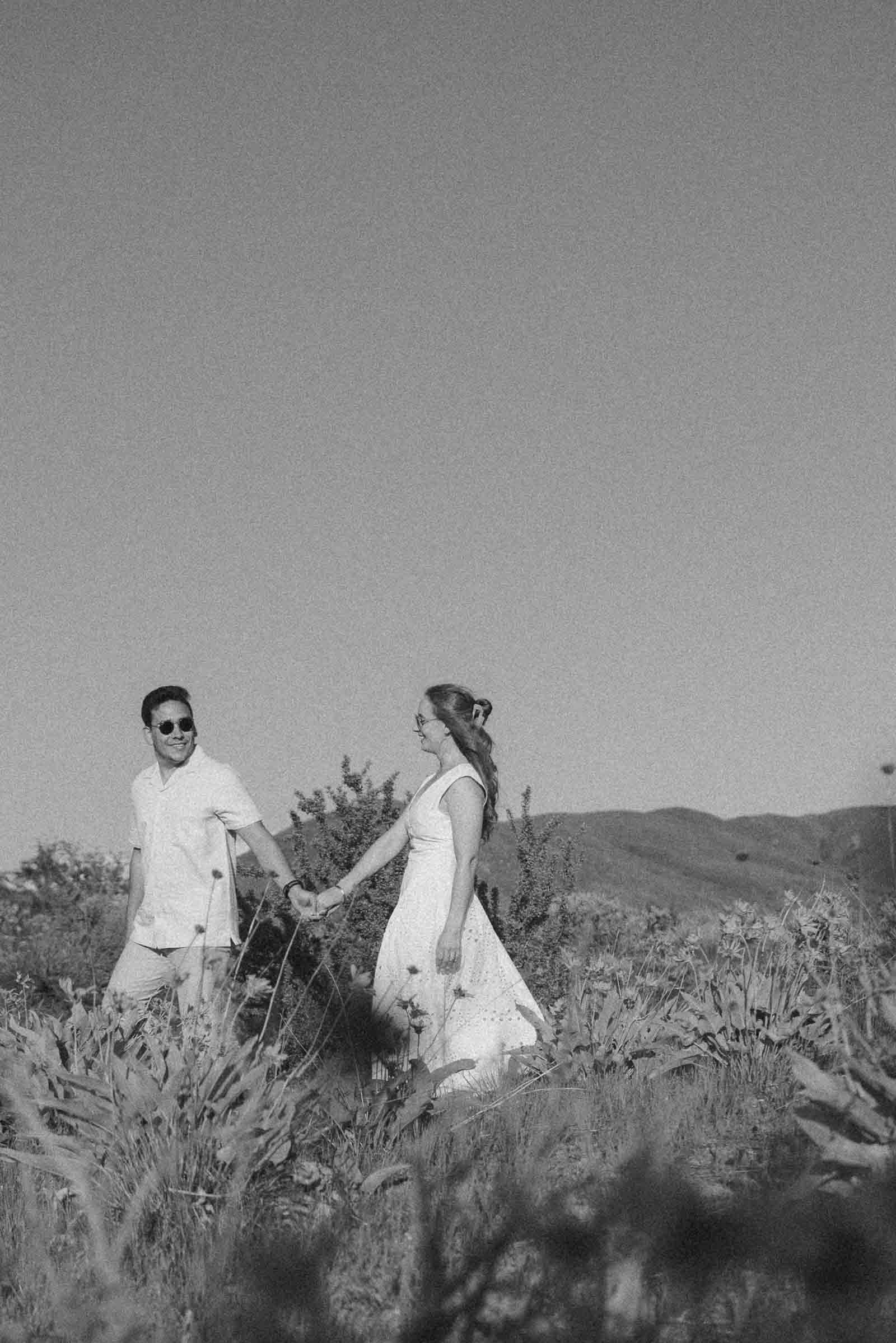  A couple in light clothing holds hands and walks through a field with wildflowers, smiling at each other. Hills and a clear sky are in the background. The photo is in black and white. 