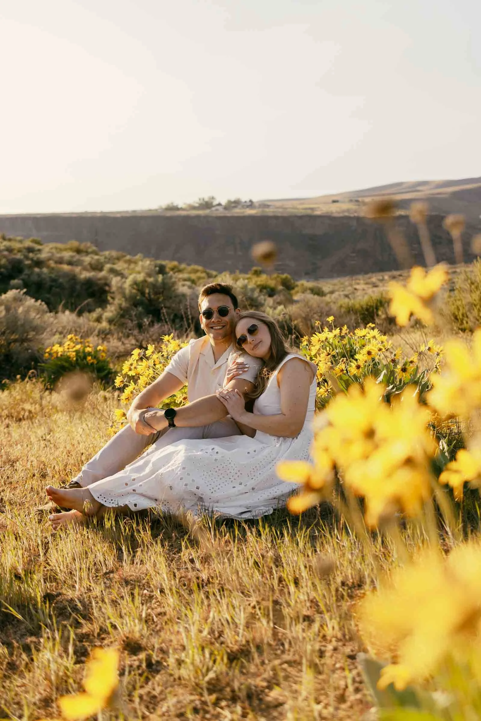  A couple sits together in a field of yellow wildflowers on a sunny day, with hills and greenery in the background. The woman wears a white dress and sunglasses, while the man is dressed casually and also wears sunglasses. 