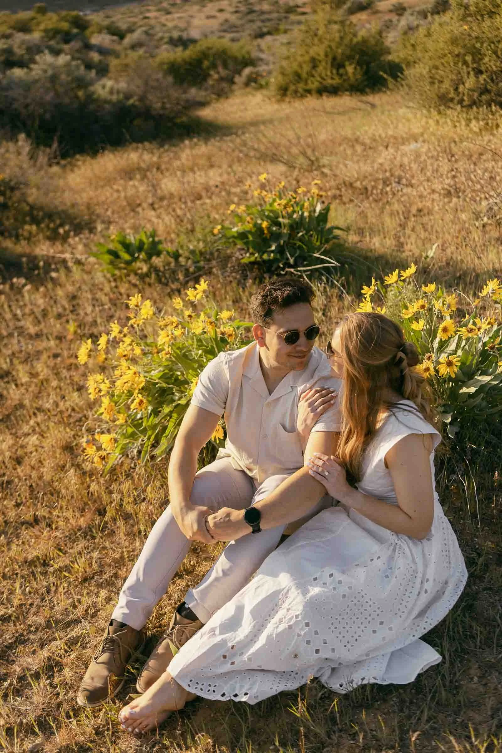  A couple sits together on dry grass in a sunlit field, surrounded by yellow flowers. The man wears sunglasses and light clothing, while the woman wears a white dress. They smile and hold each other affectionately. 