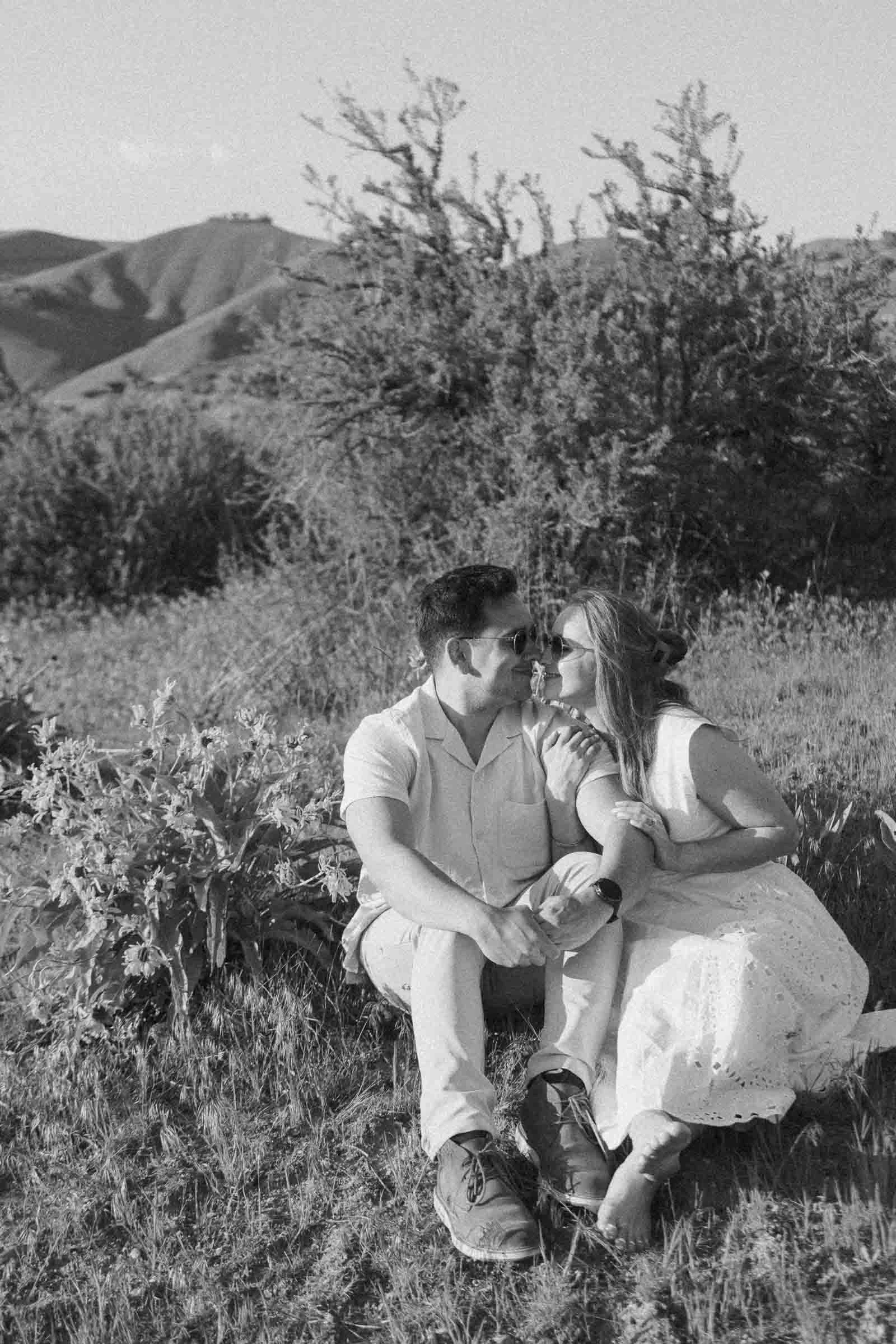  A couple sits in the grass, smiling at each other and touching noses, surrounded by bushes and hills in the background on a sunny day. The image is in black and white. 