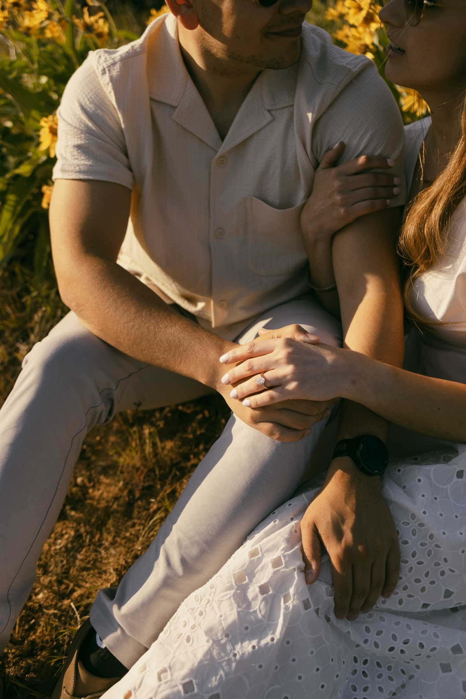  A couple sitting close together outdoors, holding hands. The woman rests her hand on the man's arm, and they are both dressed in light-colored clothing, surrounded by yellow flowers and sunlight. 
