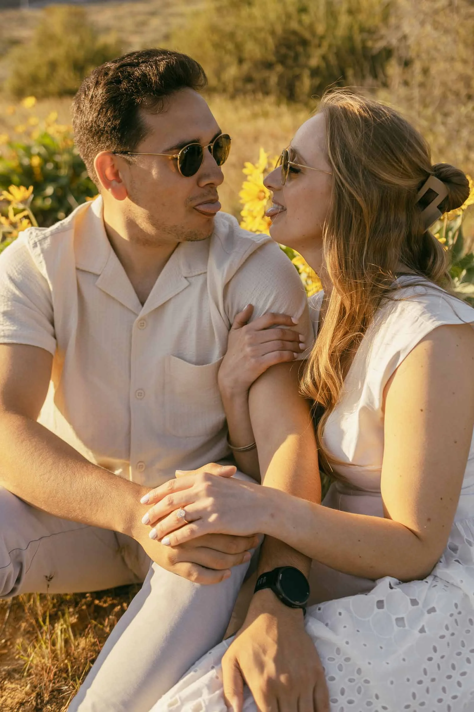  A man and woman sit close together outdoors, holding hands and smiling at each other while wearing sunglasses. They are surrounded by yellow flowers and warm sunlight. 