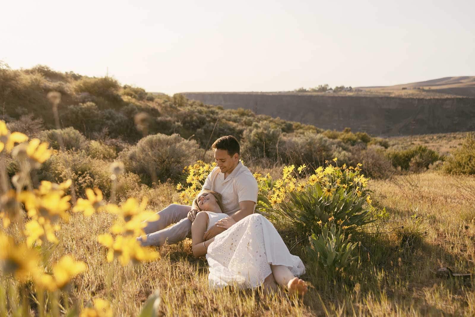  A man and woman sit together in a wildflower meadow on a sunny day, yellow blooms surrounding them on all sides. She leans into him with her head resting on his chest while he holds her close, both relaxed, with gentle hills and warm light behind th
