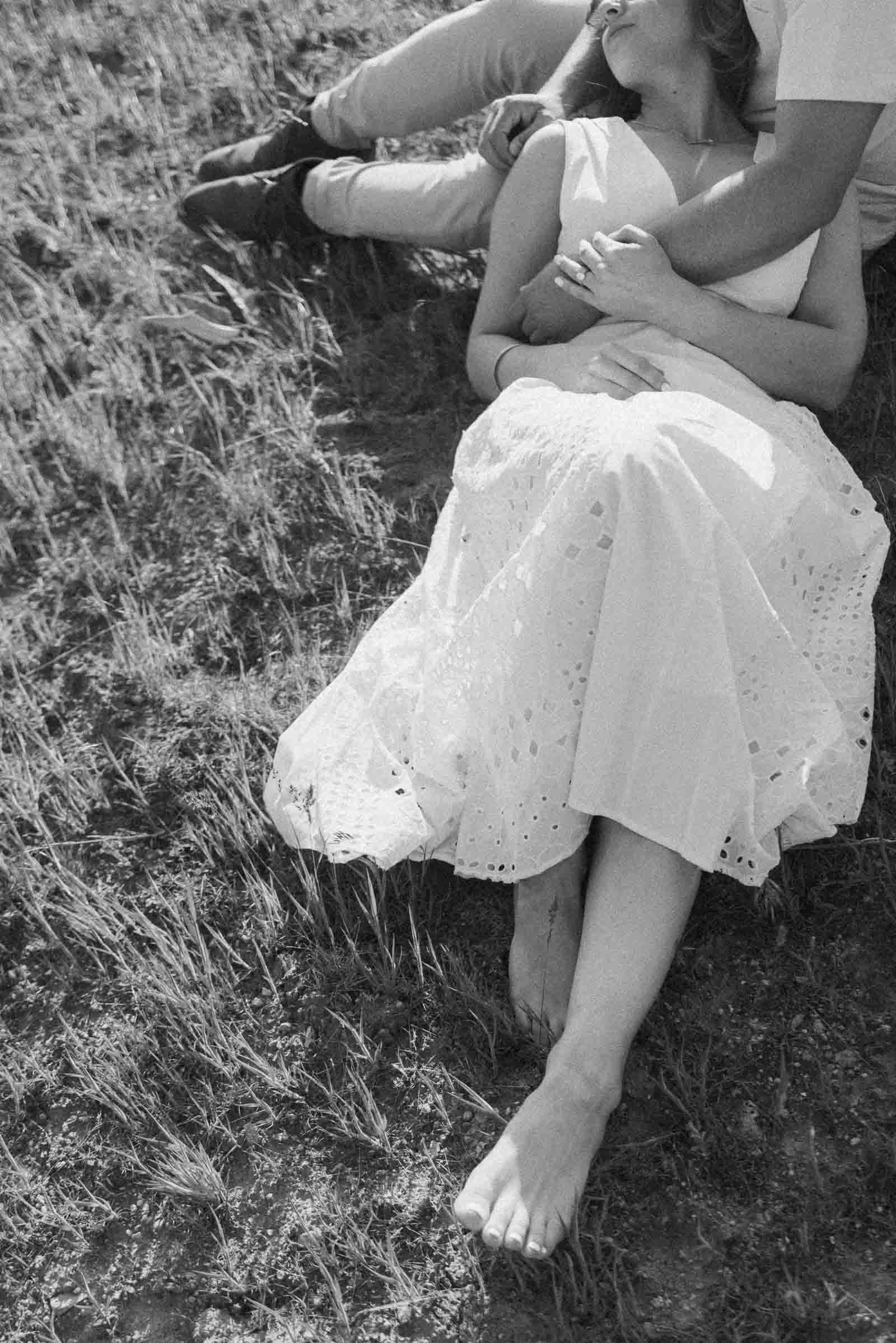  A black-and-white photo shows a woman in a white dress lying barefoot on grass, resting her head on someone’s lap. The person’s arms are gently wrapped around her as they sit close together outdoors. 