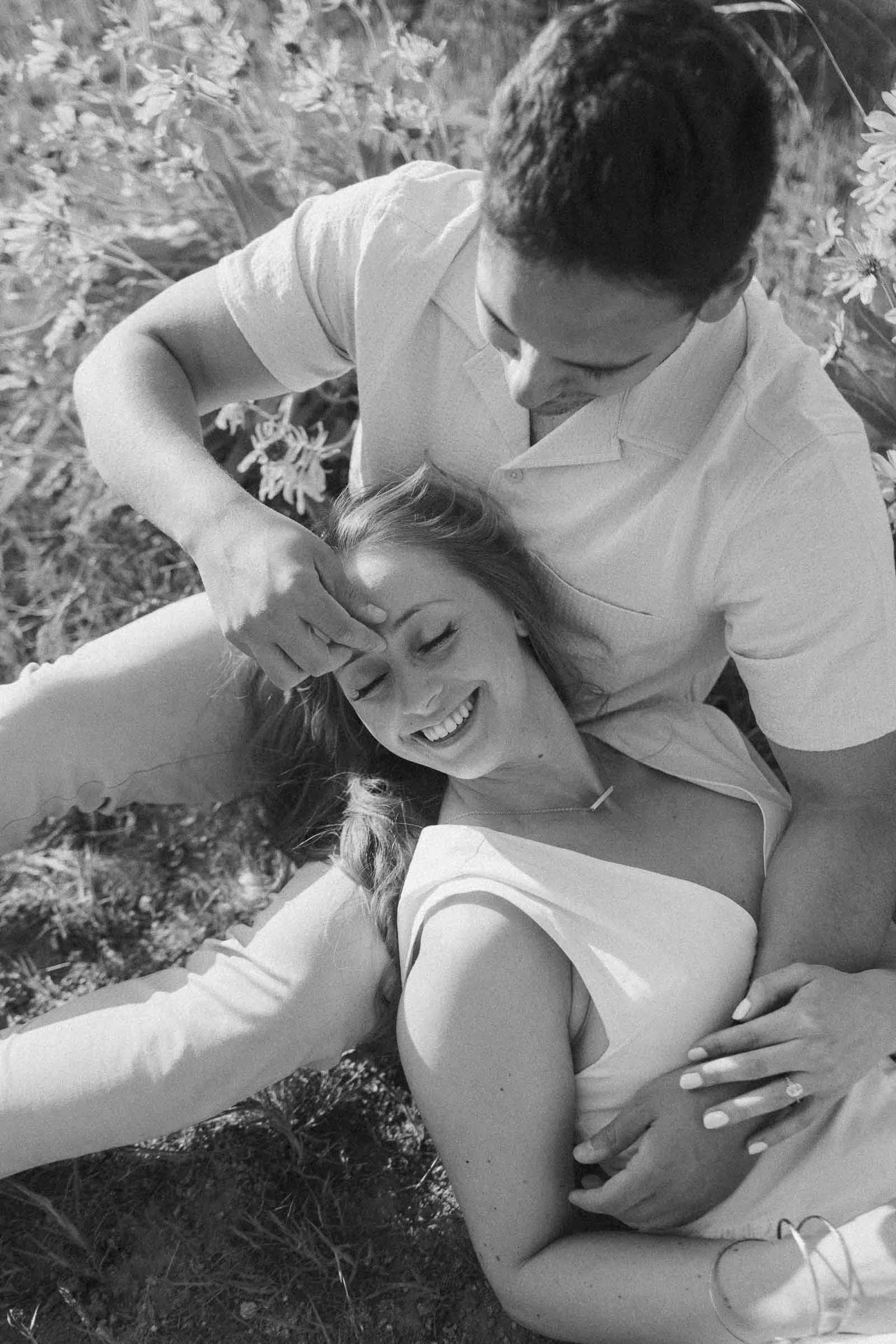  A black and white photo of a smiling couple sitting on the grass. The woman lies with her head on the man's lap, eyes closed, as he gently touches her forehead. Both appear relaxed and happy, surrounded by plants. 