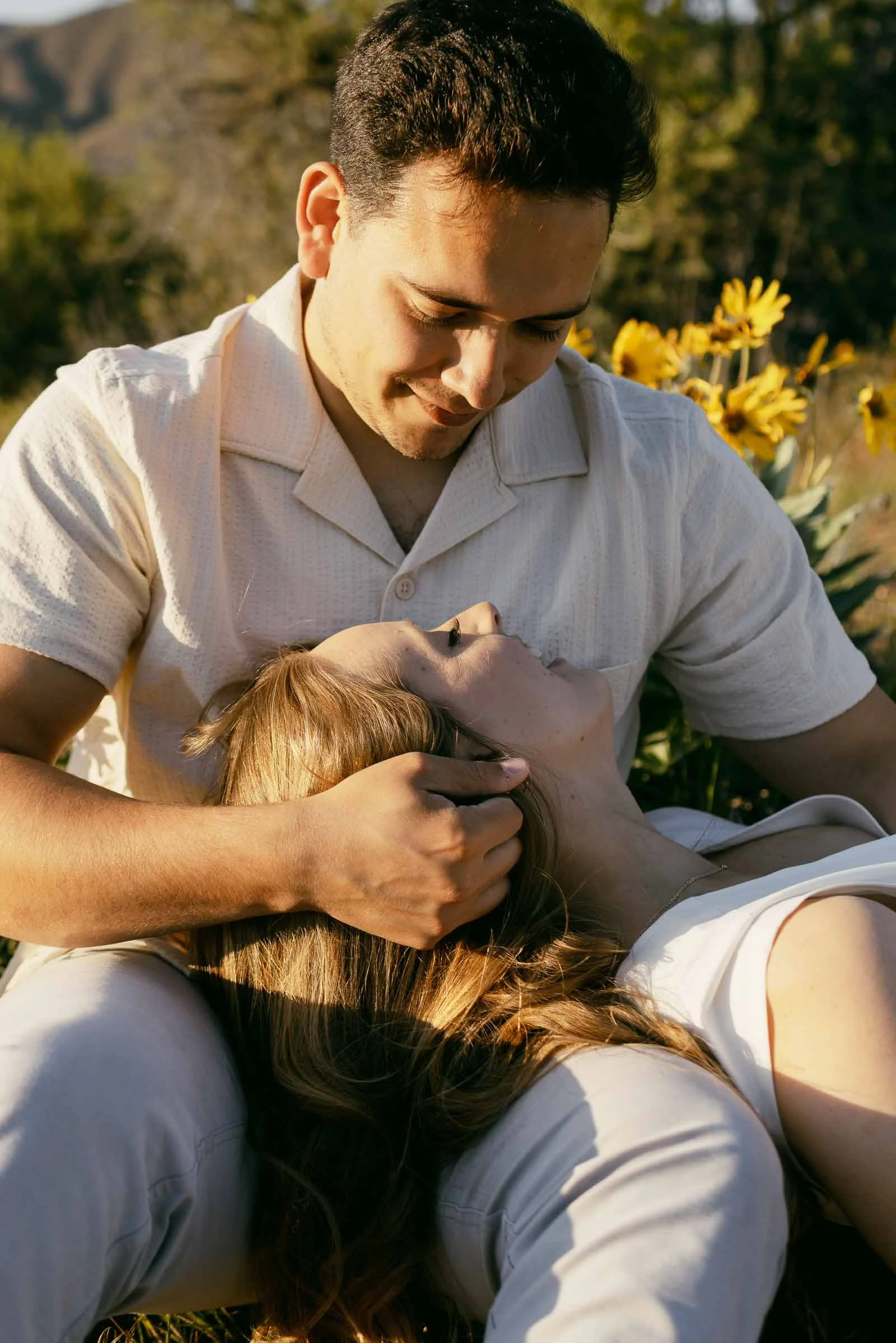  A man sitting outdoors gently cradles a woman’s head in his lap while smiling down at her. They are surrounded by green foliage and yellow flowers, and sunlight softly illuminates their faces. 