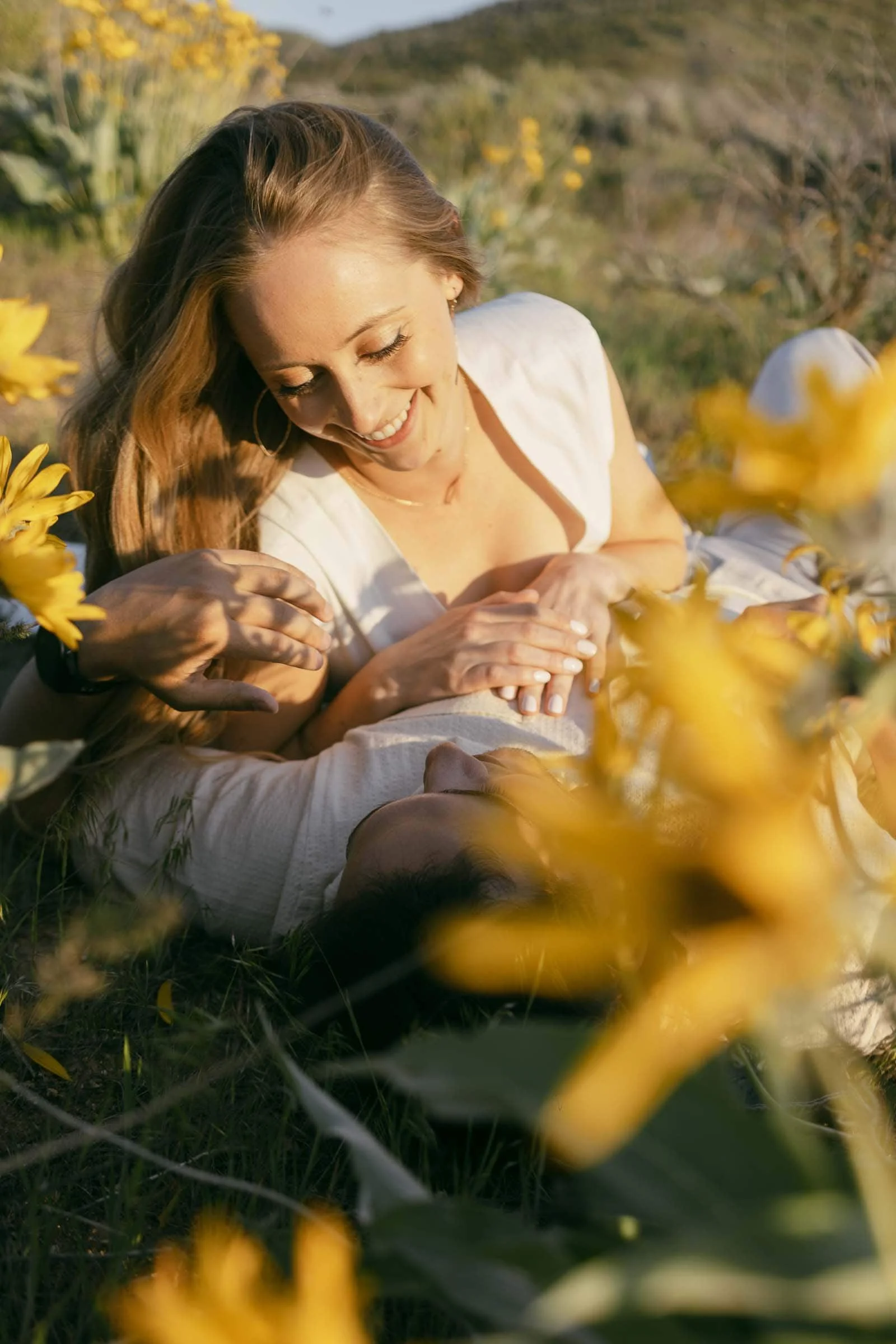  A smiling woman lies in a field of yellow flowers, her face lit by sunlight, holding the hand of someone lying next to her. The blossoms around them are bright and in full bloom. 
