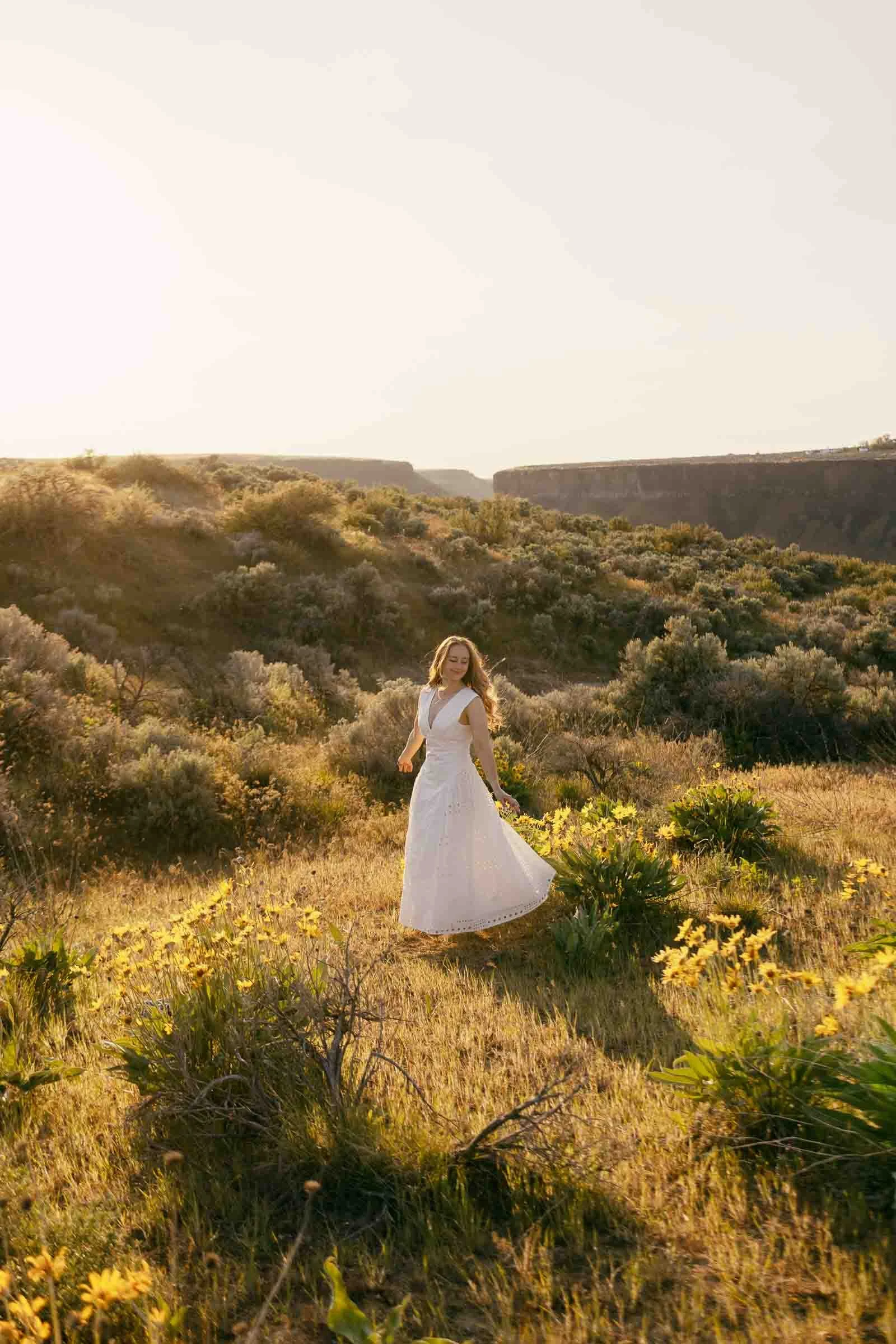  A woman in a white dress stands in a sunlit, grassy field with yellow wildflowers, surrounded by green hills and shrubs, under a bright, clear sky. 