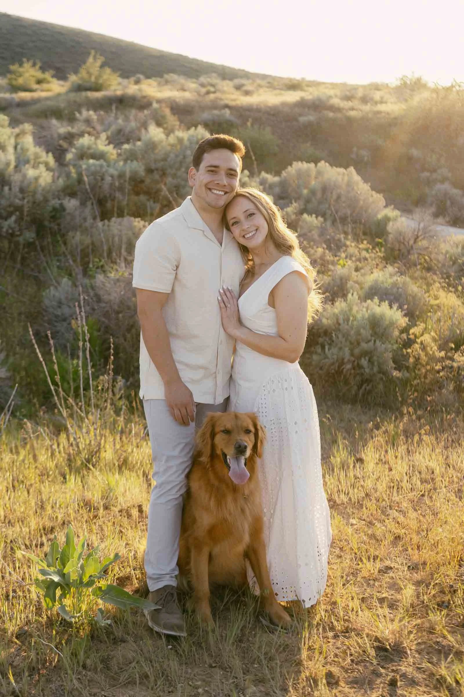  A smiling couple stands close together in a sunlit field, with a golden retriever sitting in front of them. The woman wears a white dress, and the man wears a light shirt and pants. Shrubs and hills are in the background. 