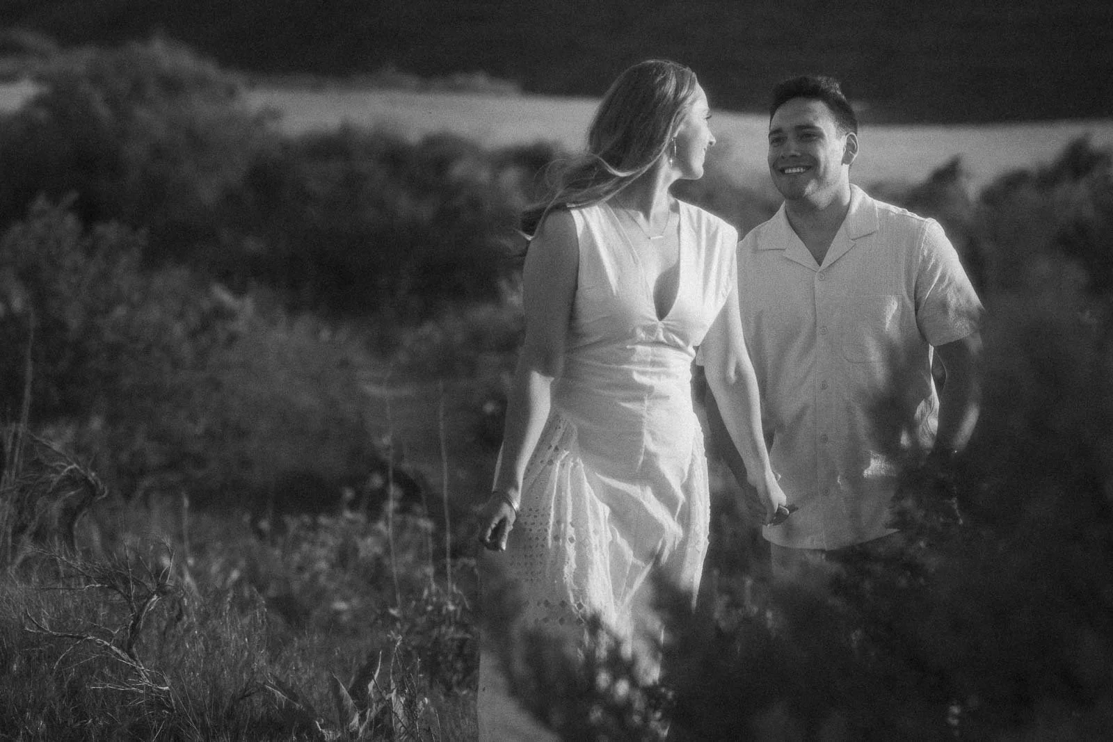 A black and white image of two people holding hands as they walk through a sun-filled grassy field, both smiling and glancing toward each other. The soft light and open setting give the photo a quiet, unhurried feeling.