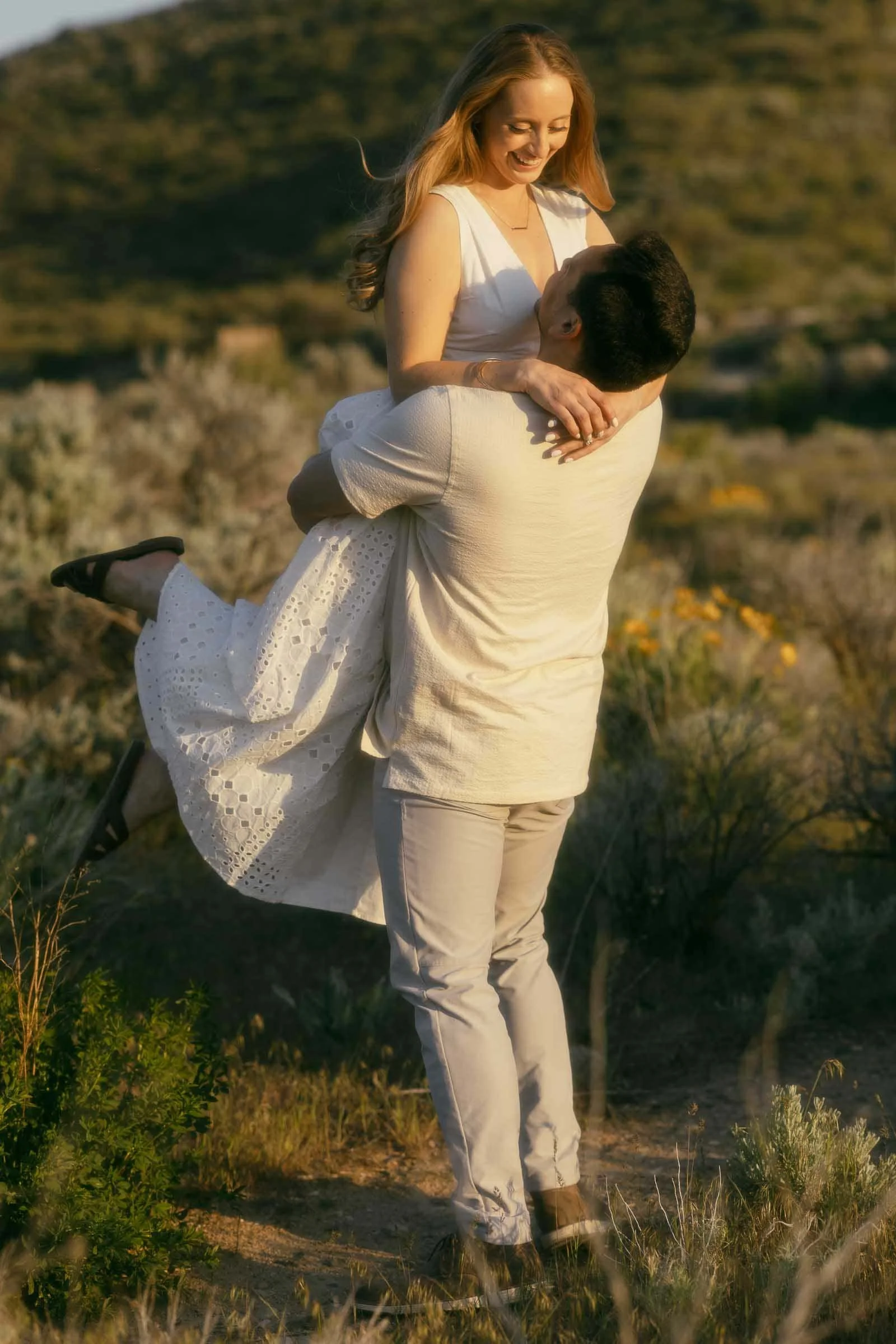  A man lifts a smiling woman in a white dress outdoors in a sunlit, grassy area, with green hills in the background. The couple looks happy and affectionate. 