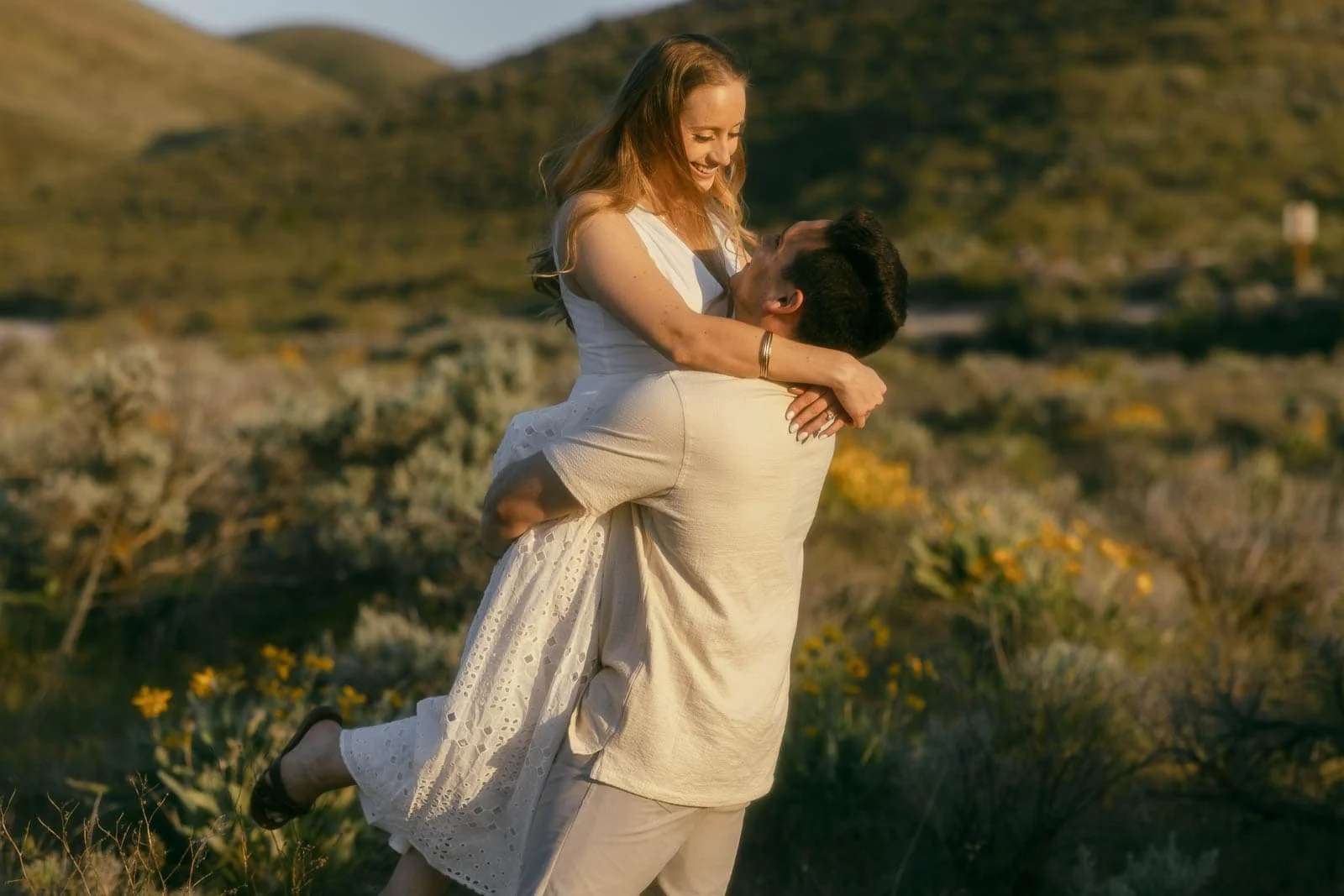  A man lifts a woman in a white dress as they smile at each other, surrounded by sunlit greenery and wildflowers in a hilly outdoor setting. 