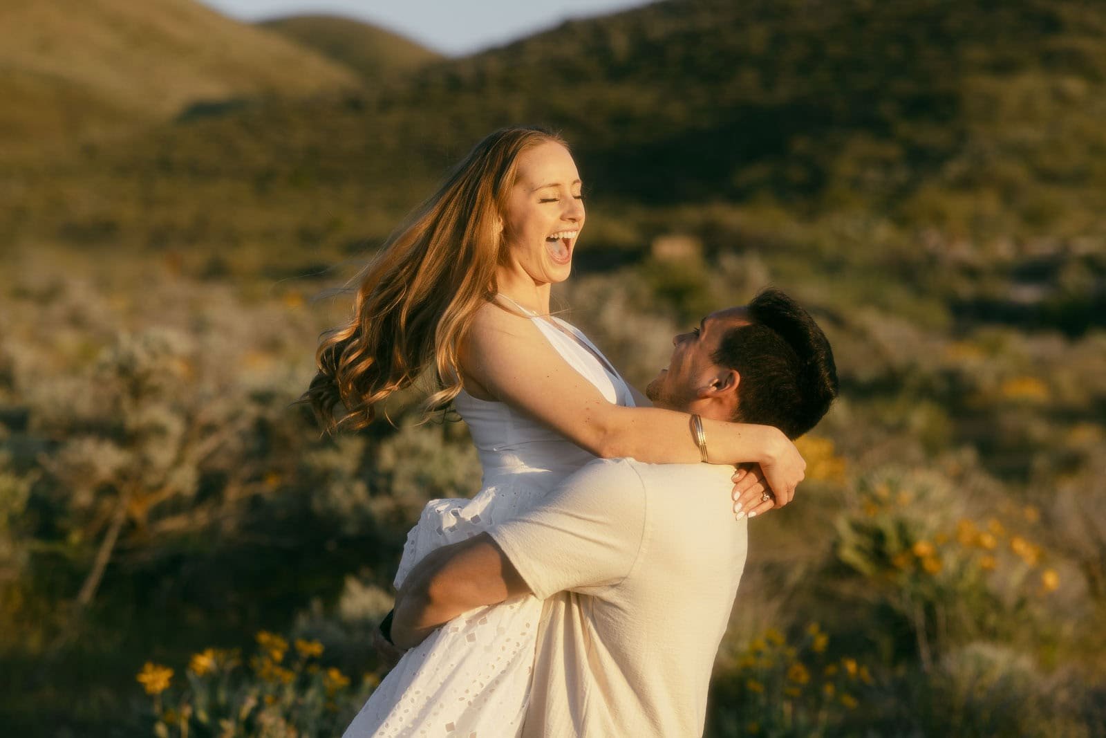  A man lifts a smiling woman in a white dress under the warm sunlight in a scenic field with hills and wildflowers in the background. 