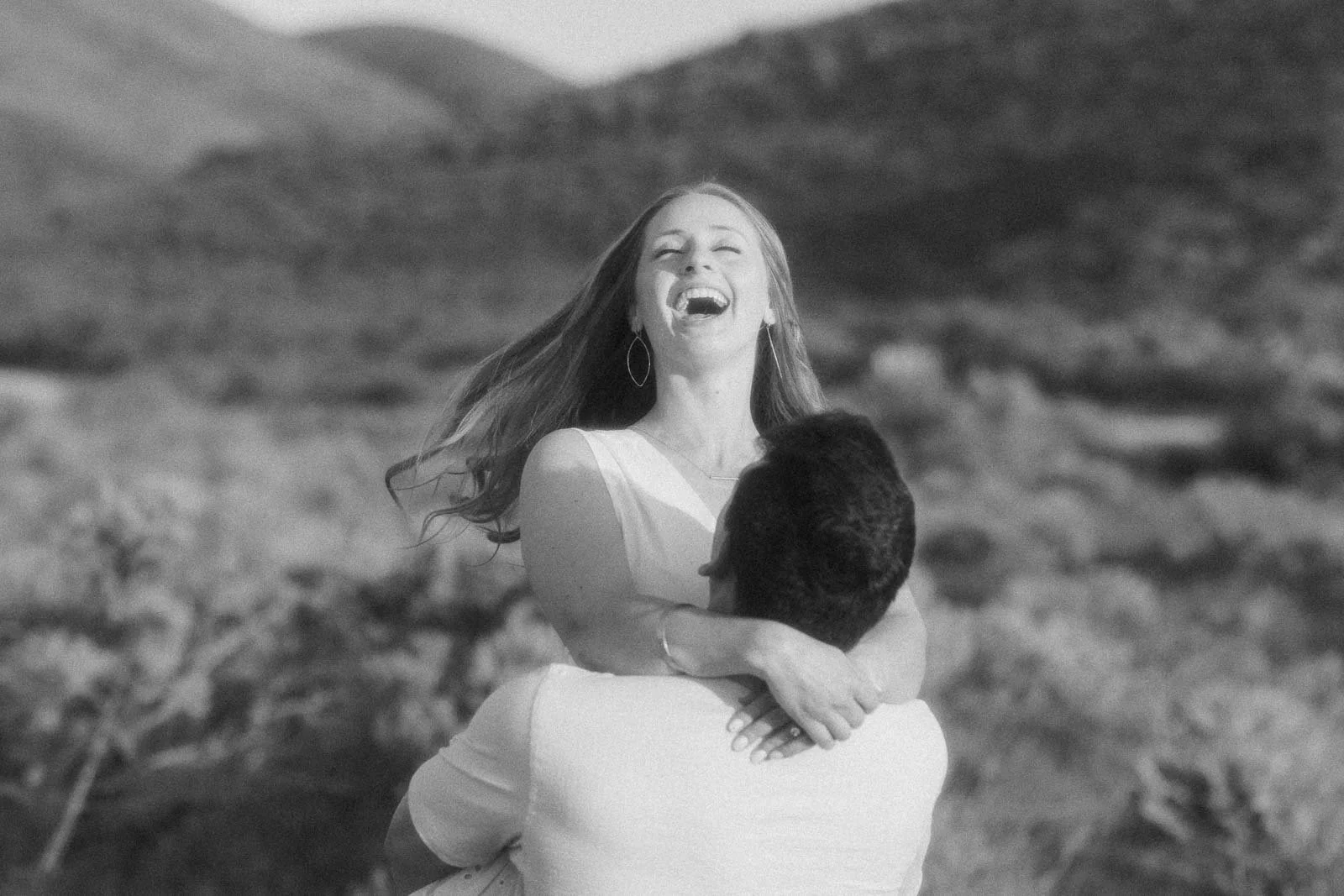  A joyful woman with long hair laughs while being lifted and embraced by a man outdoors in a sunlit, natural setting with blurred hills and greenery in the background. The image is in black and white. 