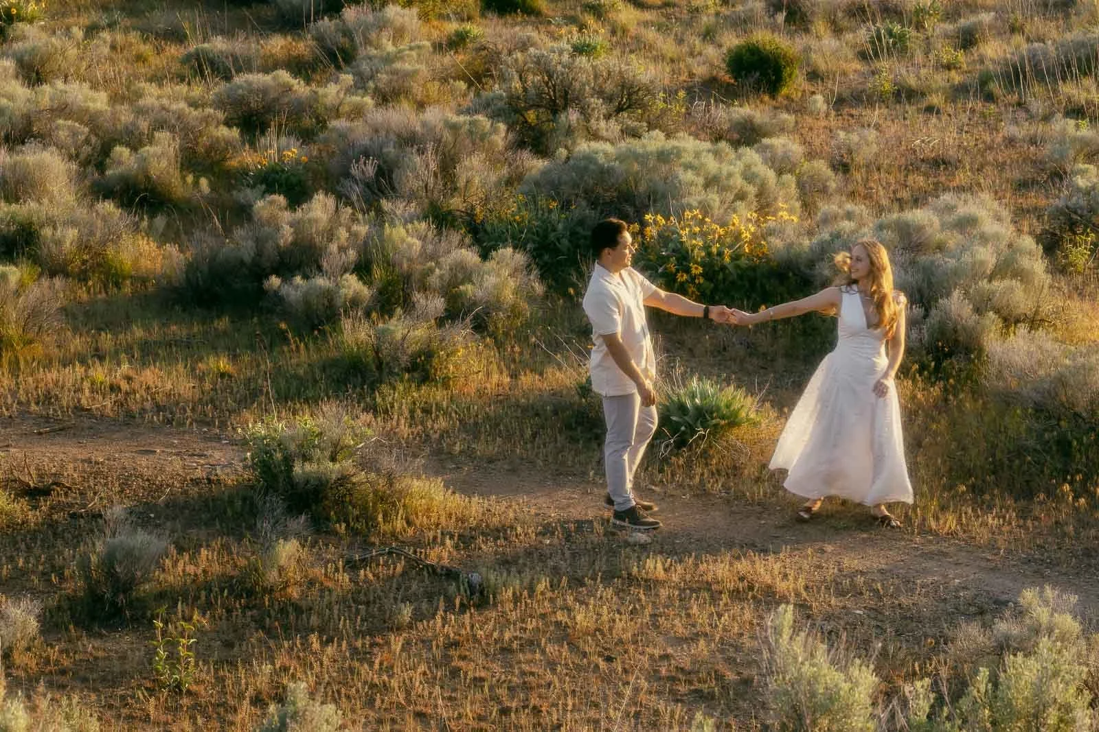  Two people in light-colored clothing hold hands and spin together on a sun-drenched dirt trail, dry grass and desert brush stretching out around them on both sides. 