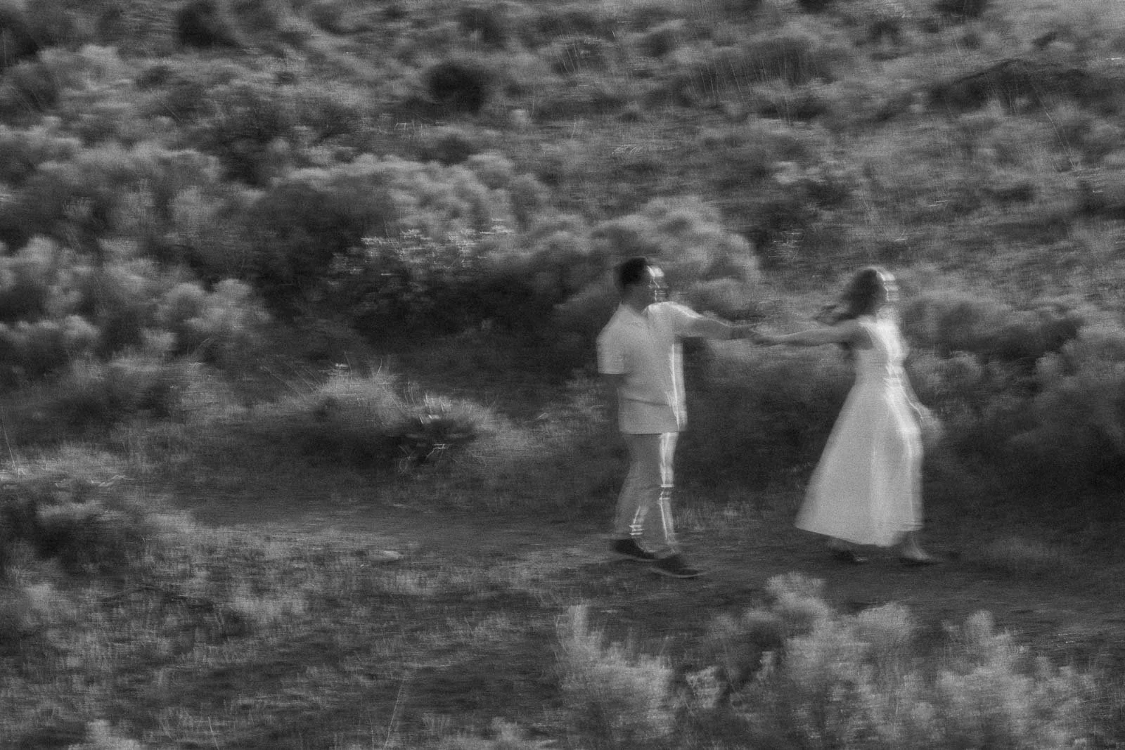  A motion-blurred black and white image of a couple dancing on a dirt path outdoors, surrounded by wild grass and low shrubs. The woman's long white dress sweeps as they move, and the man is dressed in light clothing beside her. 