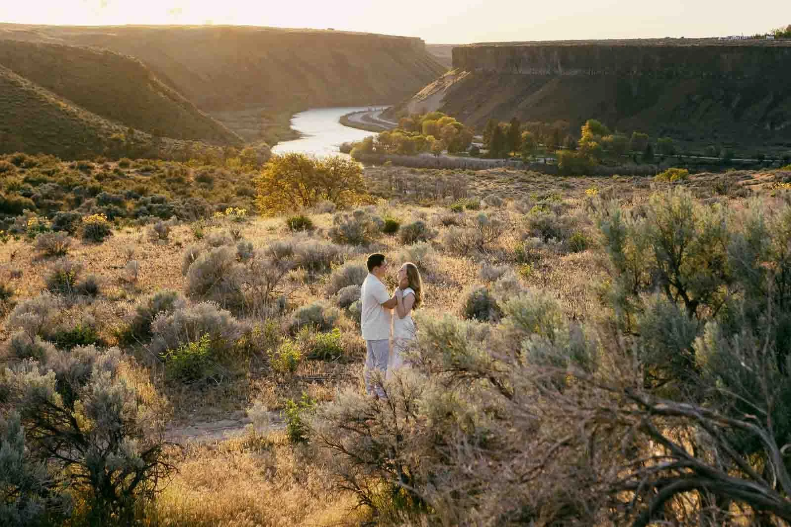  A couple stands close together in a sunlit, grassy landscape with shrubs and hills. A river winds through the background, surrounded by cliffs, under a golden sky at sunset. 