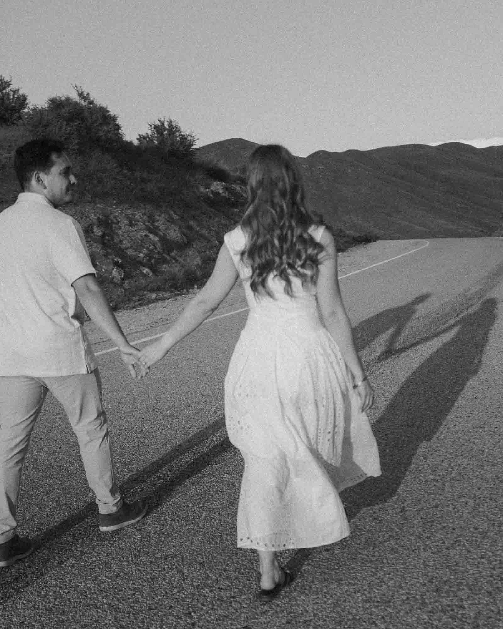  A couple holds hands and walks down an empty road in a rural, hilly area. The woman wears a long white dress, and the photo is in black and white. Their shadows stretch out behind them on the road. 