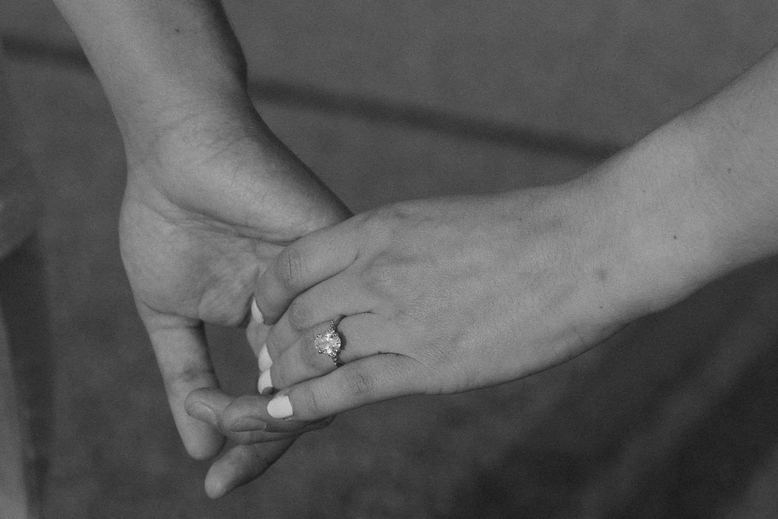  Two hands holding each other gently, with one hand wearing a sparkling engagement ring on the ring finger. The image is in black and white. 
