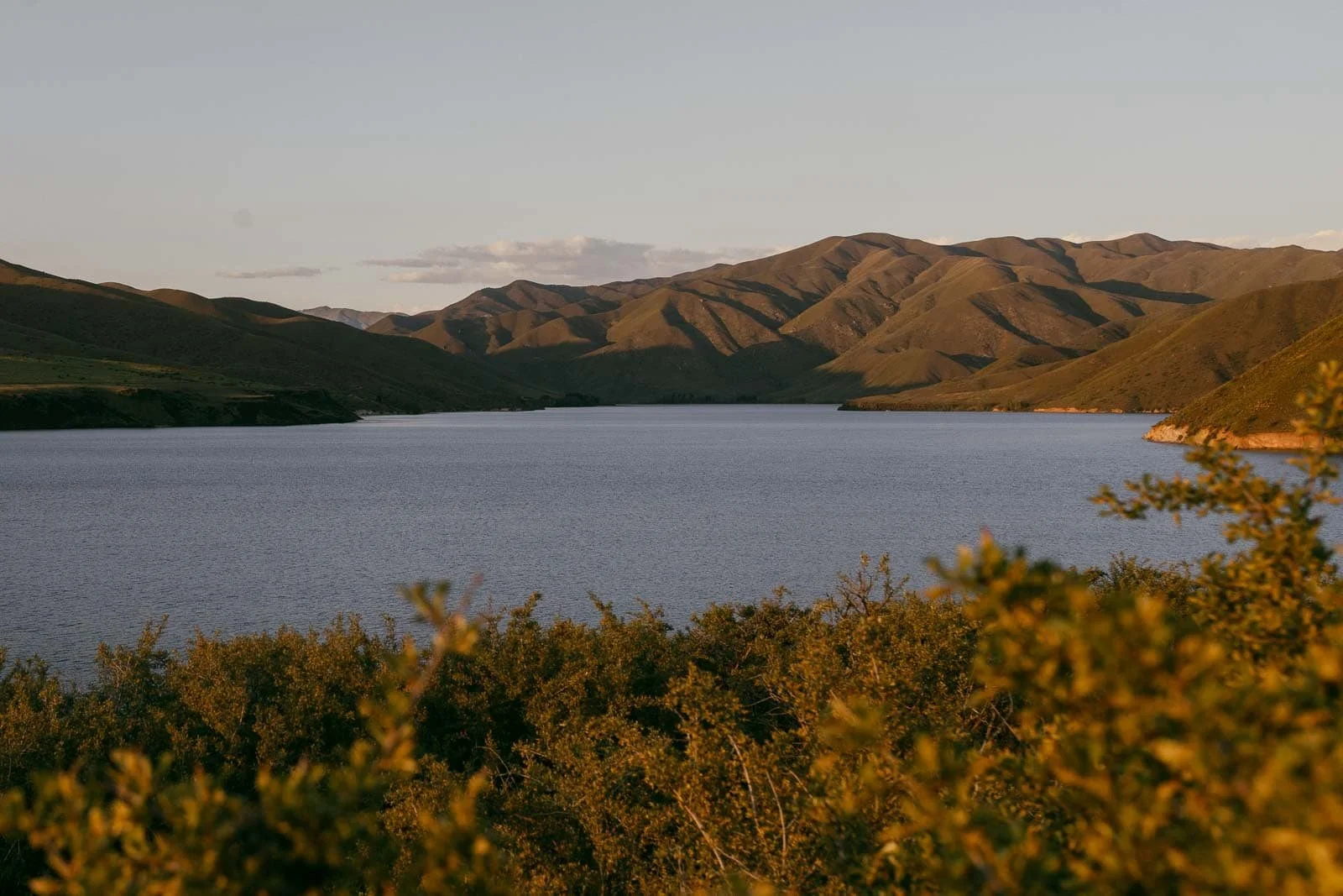  A calm lake is surrounded by green shrubs in the foreground and rolling, sunlit hills in the background under a clear sky during golden hour. 