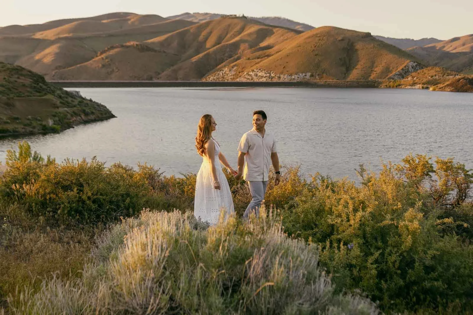 A couple holding hands and walking near a lake, surrounded by green bushes and hills under a clear sky at sunset. The woman wears a white dress, and the man is in light clothing. 