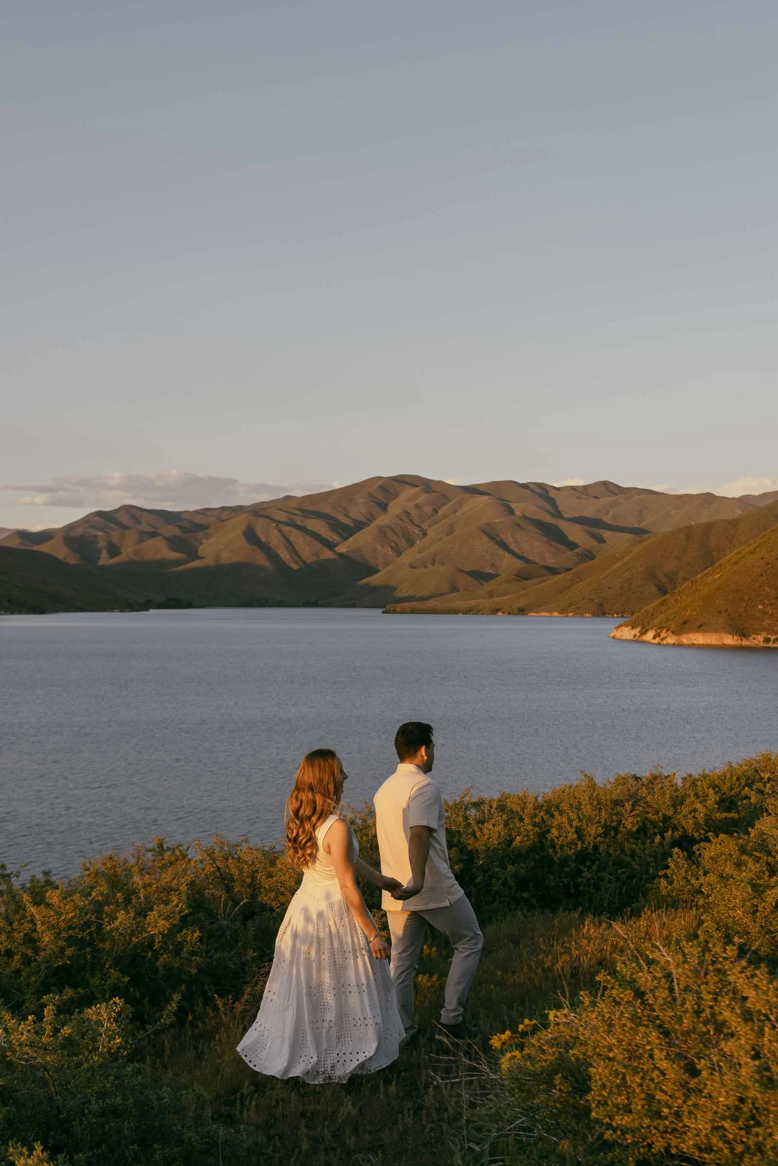  A couple stands side by side, facing a serene lake surrounded by green hills under a golden sunset sky. The woman wears a white dress, and the man wears light-colored clothes. Bushes and grass fill the foreground. 
