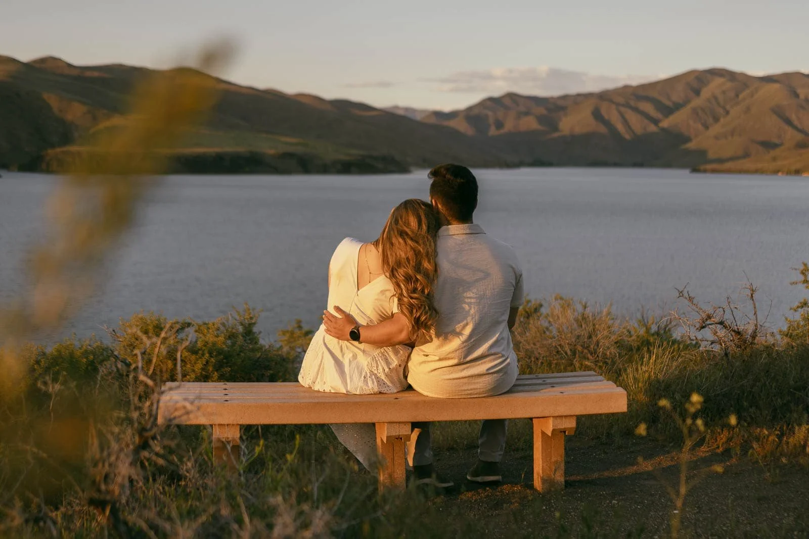  A couple sits close together on a wooden bench overlooking a calm lake and mountains at sunset, with the woman resting her head on the man's shoulder. 