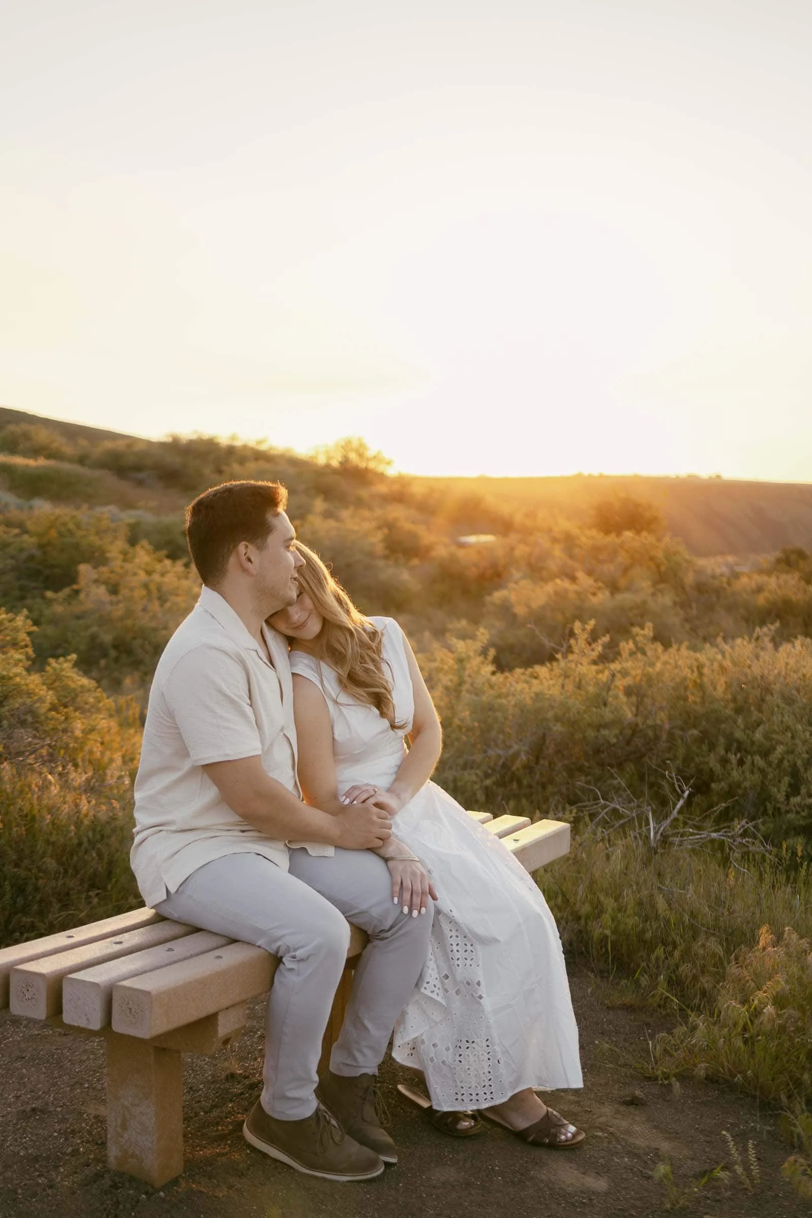  A couple sits closely on a wooden bench outdoors at sunset, surrounded by greenery. The woman rests her head on the man's shoulder as they hold hands, looking peaceful and content. 