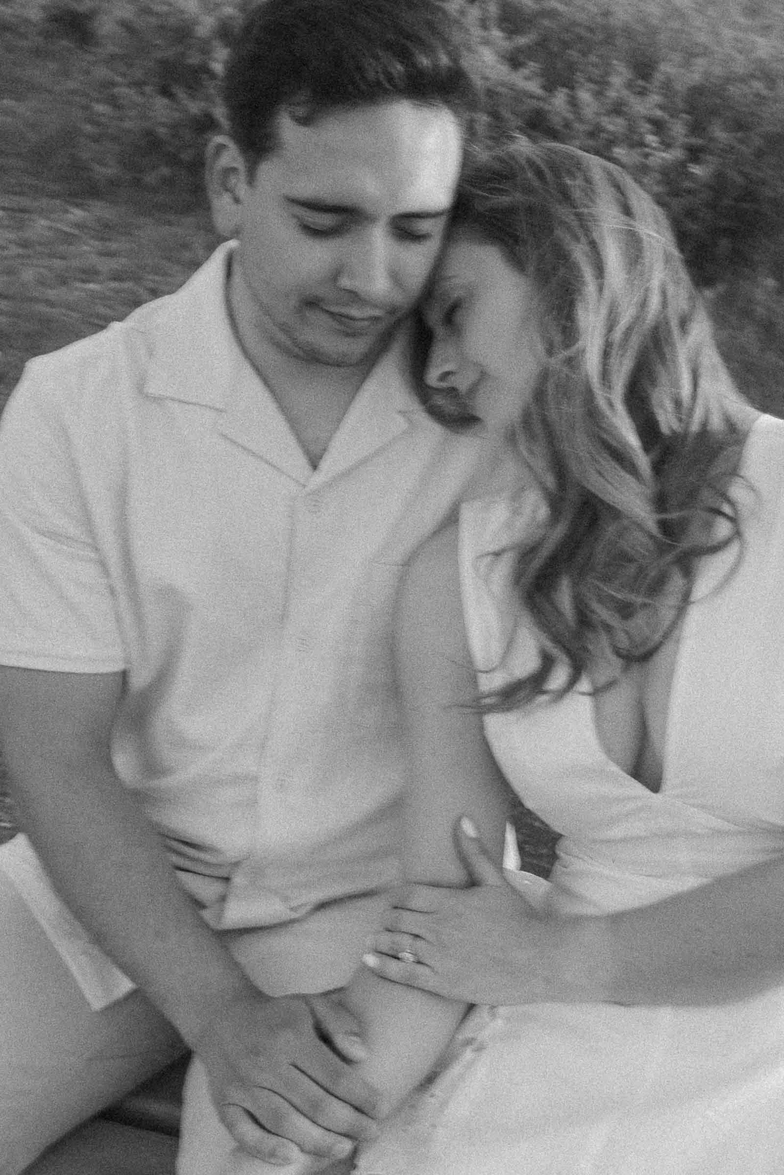  A black and white photo of a couple sitting closely together outdoors, with the woman resting her head on the man's shoulder and their hands gently touching, both appearing peaceful and affectionate. 