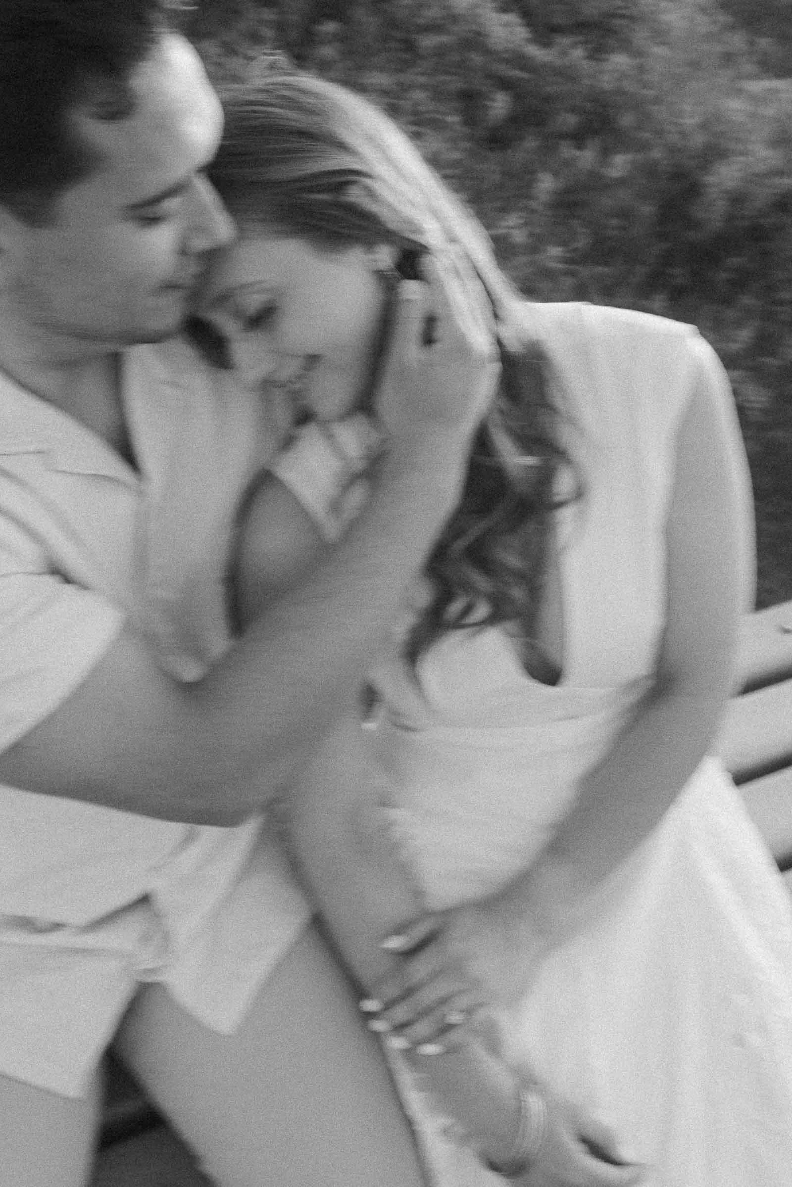  A black and white photo of a couple sitting closely together on a bench. The man gently touches the woman’s face as she leans into him, smiling softly, creating an intimate and tender moment. 