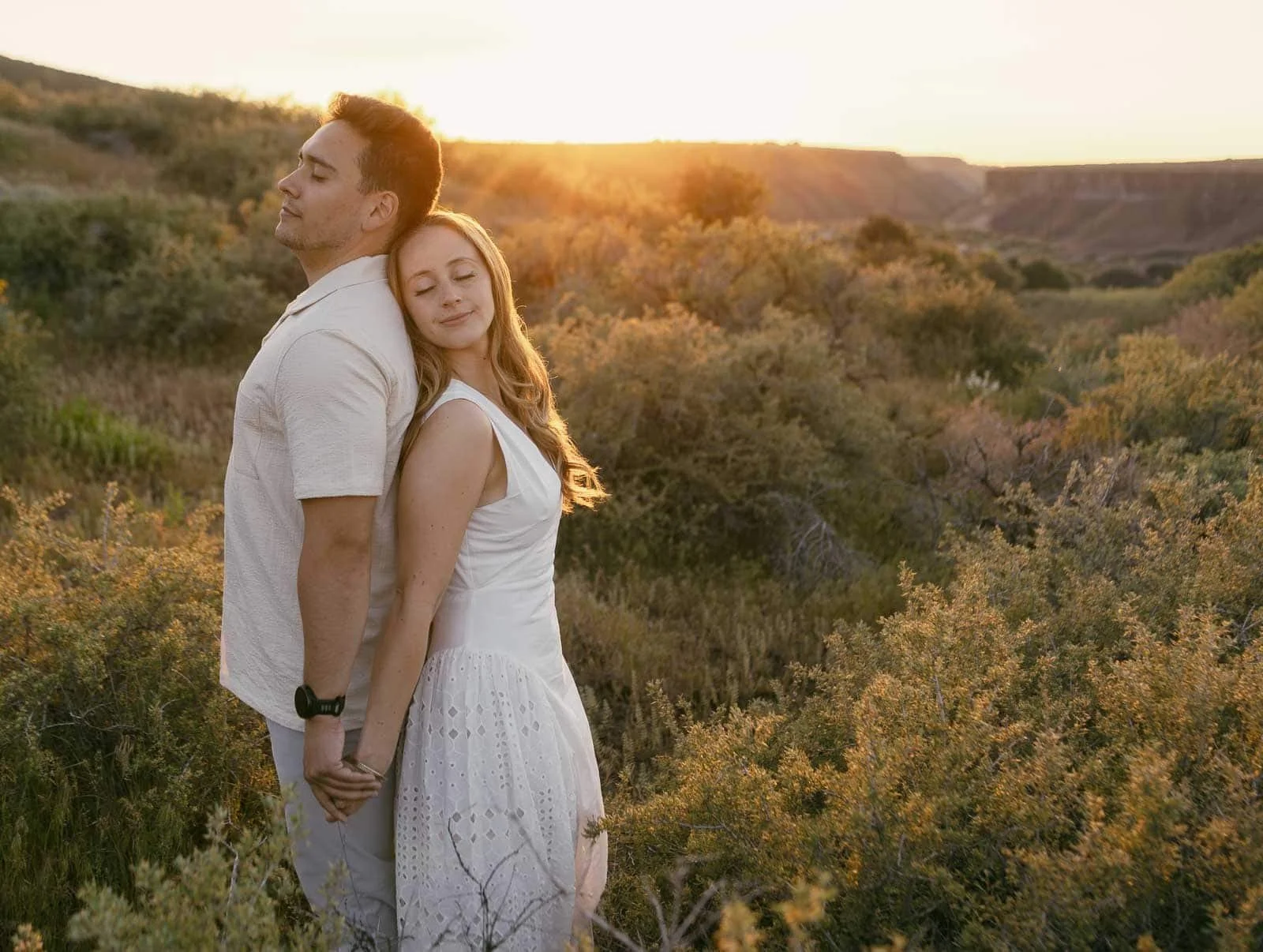 A man and woman stand back to back at sunset, hands clasped and eyes closed, both smiling. Tall grasses, dense bushes, and sun-warmed hills surround them on all sides.