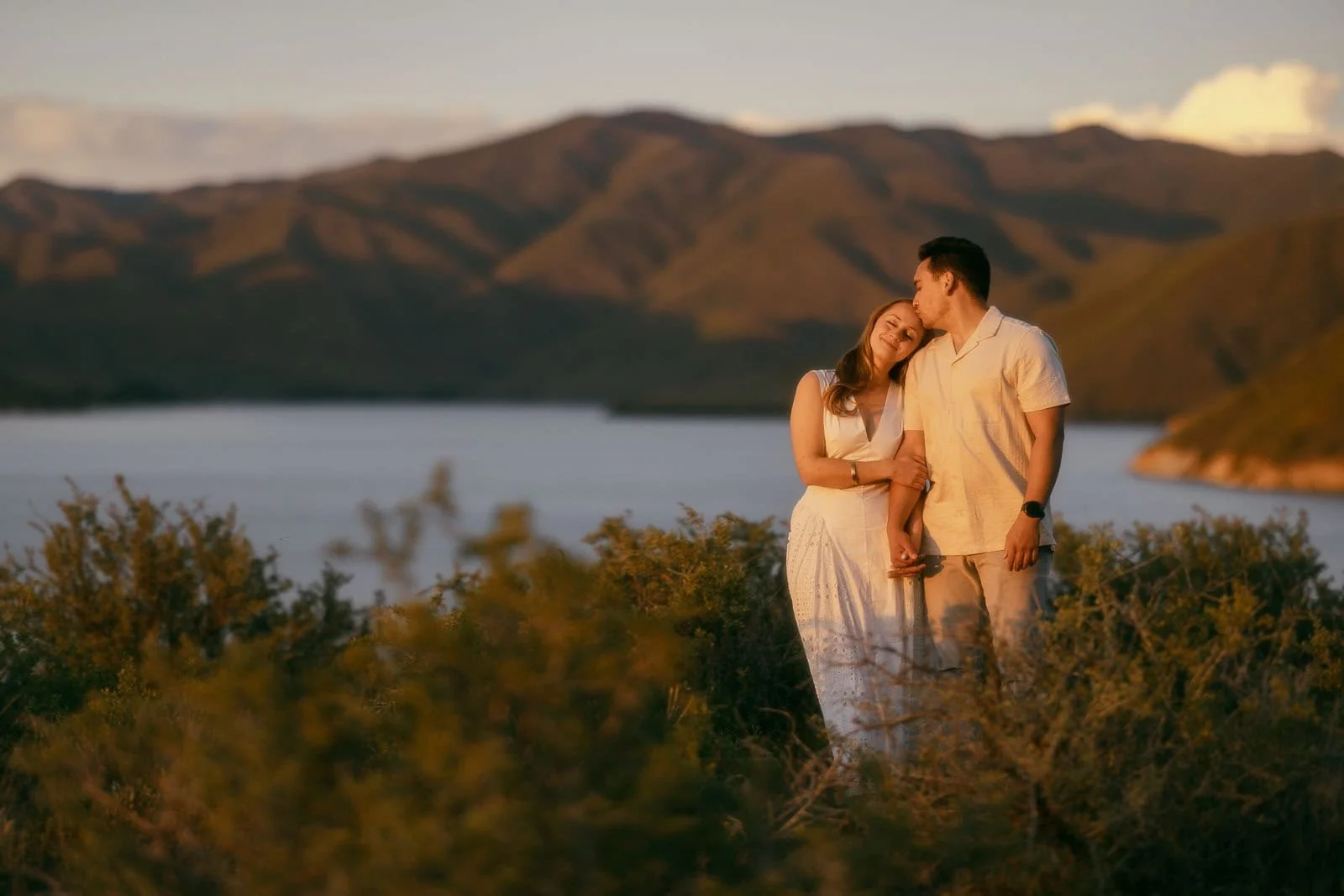  A couple stands close together outdoors, with the woman resting her head on the man's shoulder. They are surrounded by greenery, with a lake and mountains in the background under a warm, golden light. 