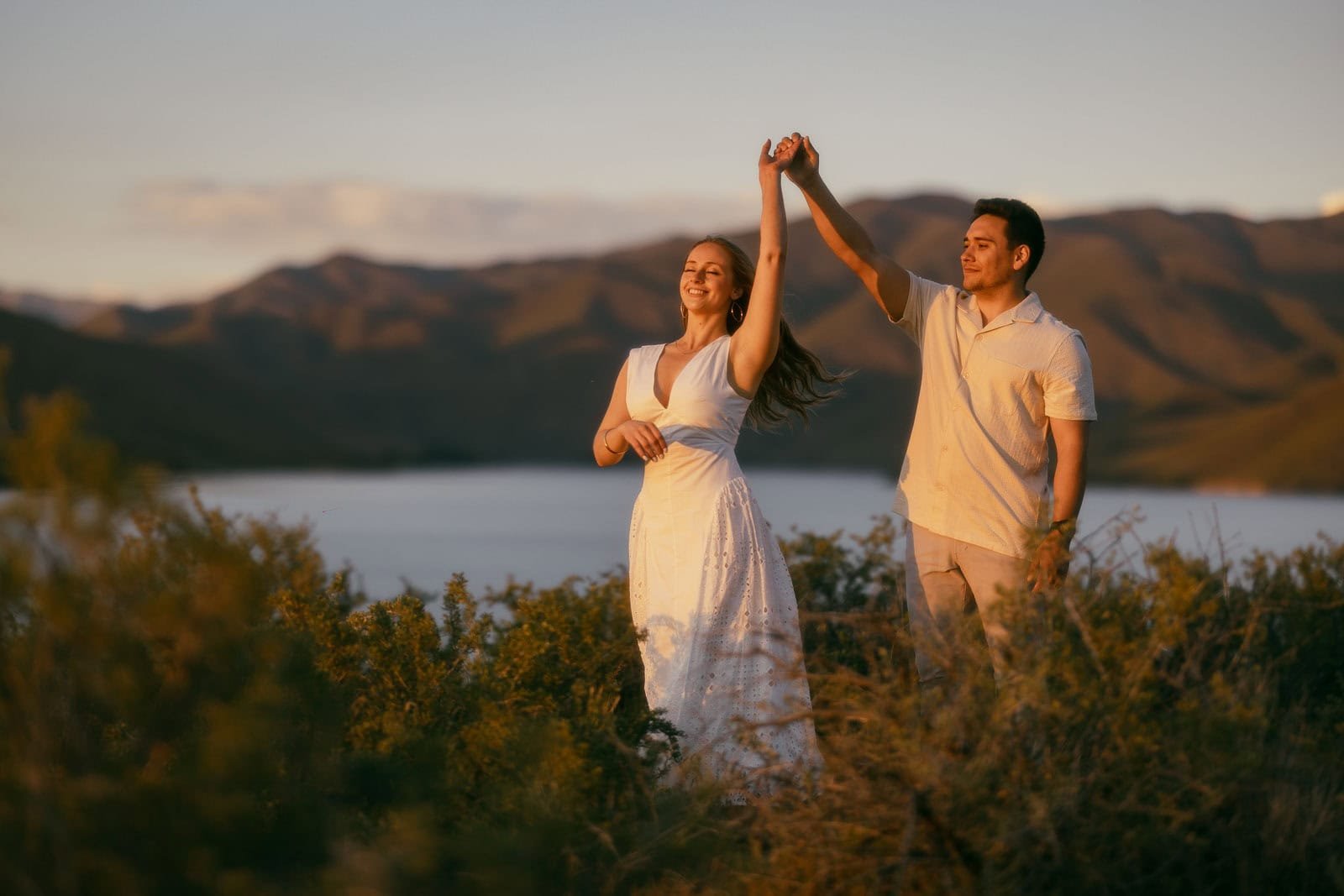 A couple dances in an open field at golden hour, a lake and mountain range visible in the distance behind them. She wears a white dress and he wears a light shirt and pants, both laughing as they move together.