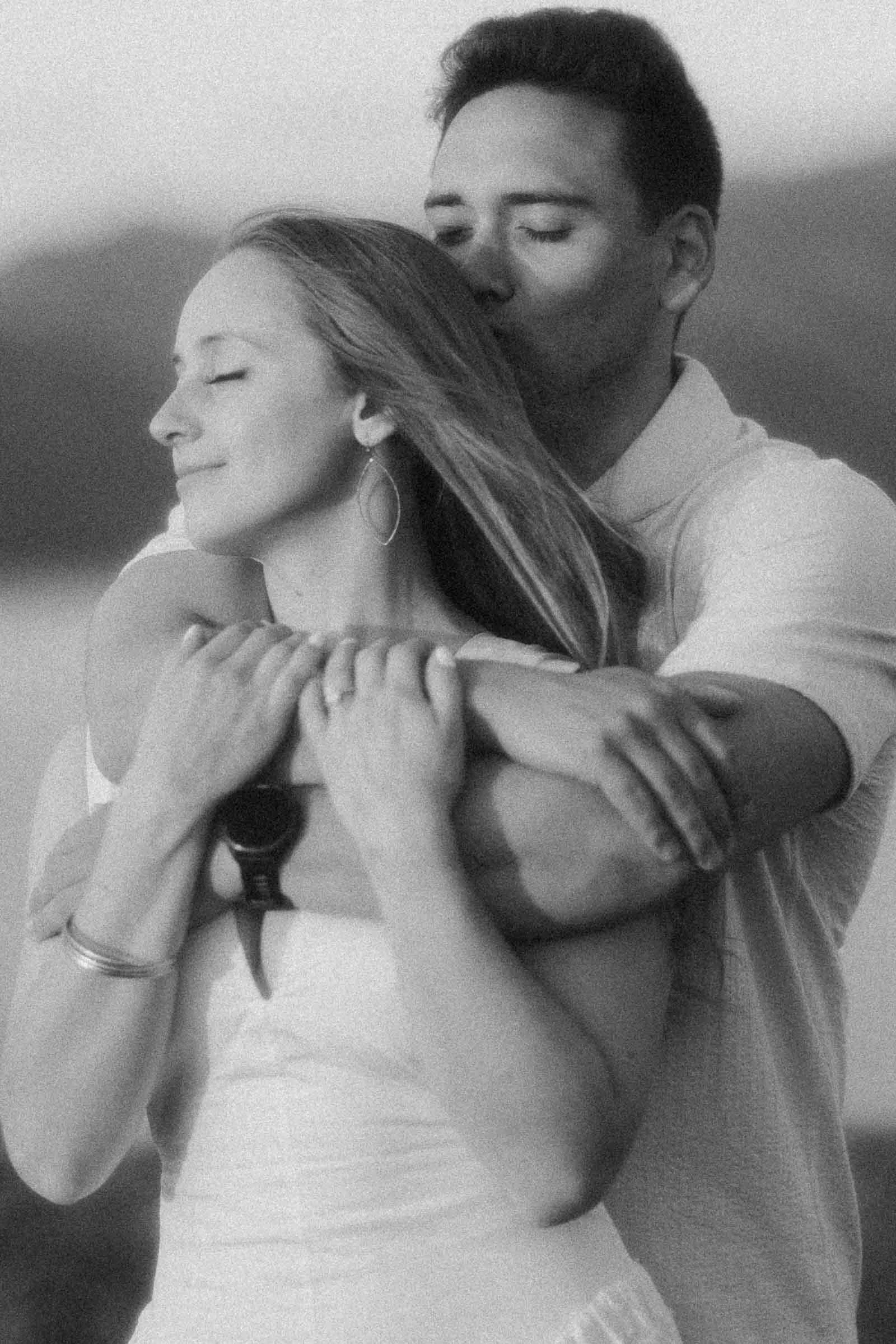  A man gently embraces a woman from behind, kissing her head as she smiles with her eyes closed. Both appear serene and content, dressed in light clothing, with a blurred natural background. 