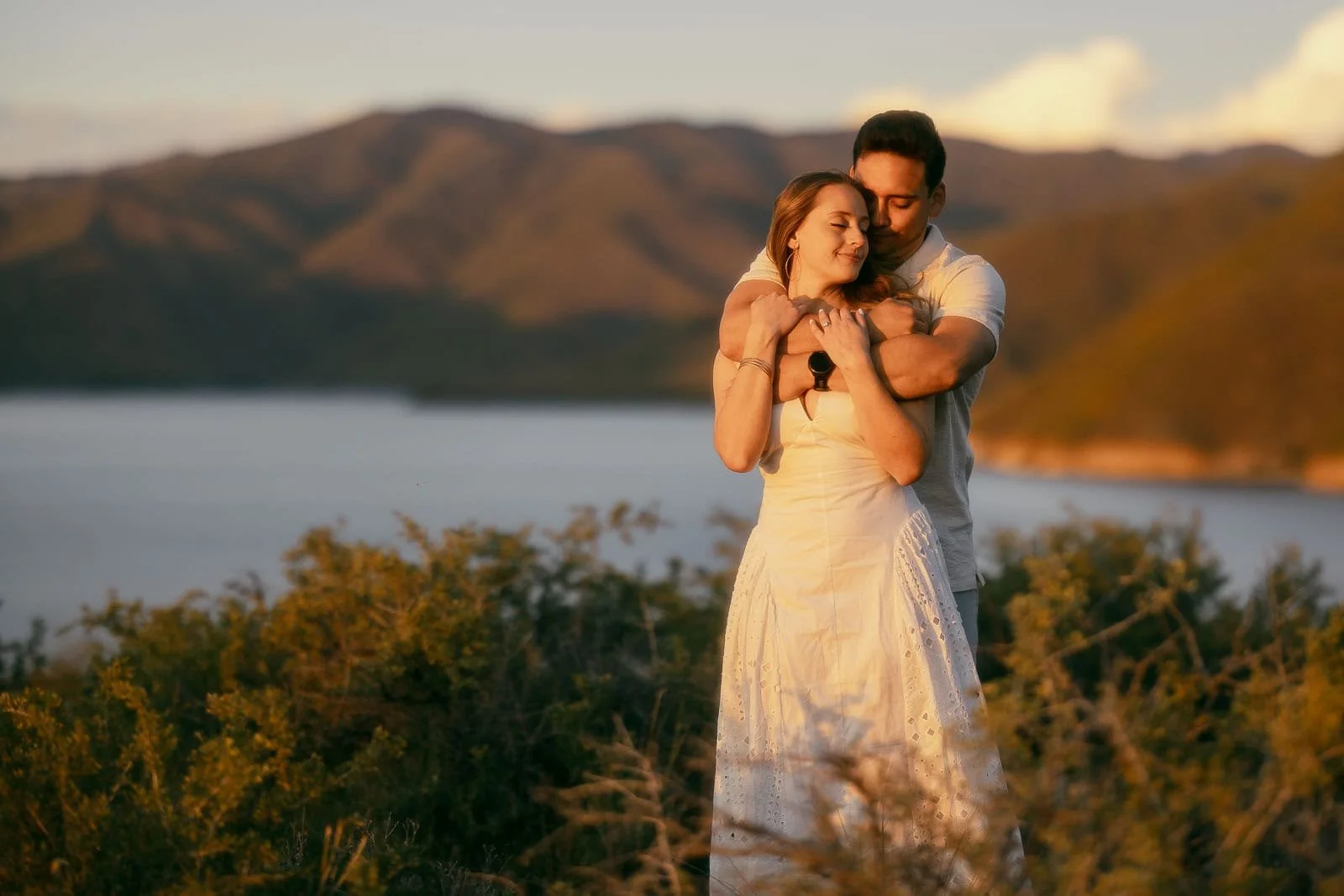  A man stands behind a woman in a white dress at sunset, arms wrapped around her as she leans back into him, eyes closed and smiling softly. A calm lake and mountain ridgeline fill the background behind them. 