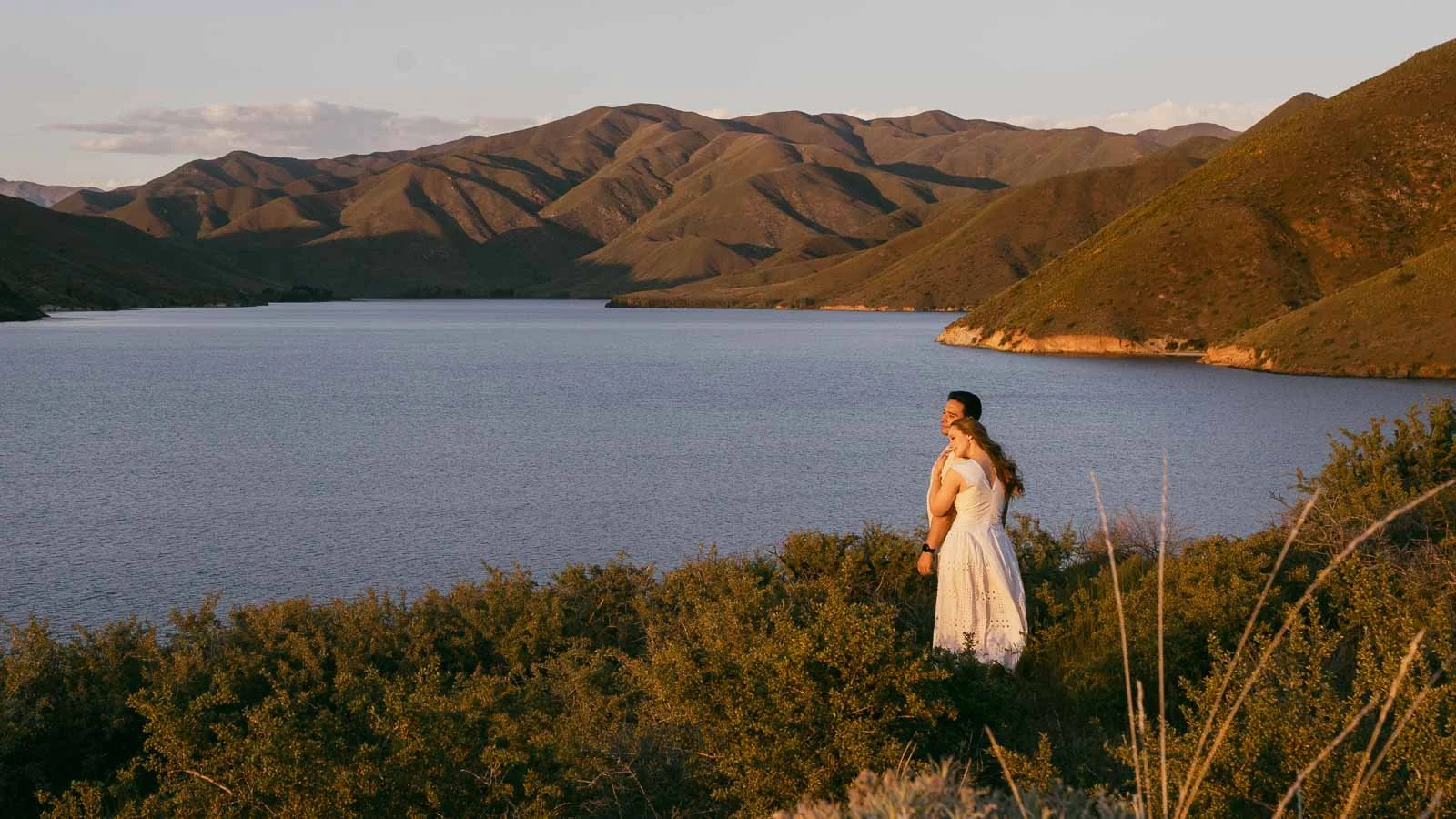  A woman in a white dress stands on a grassy hill overlooking a large lake, surrounded by green mountains under a partly cloudy sky at sunset. 