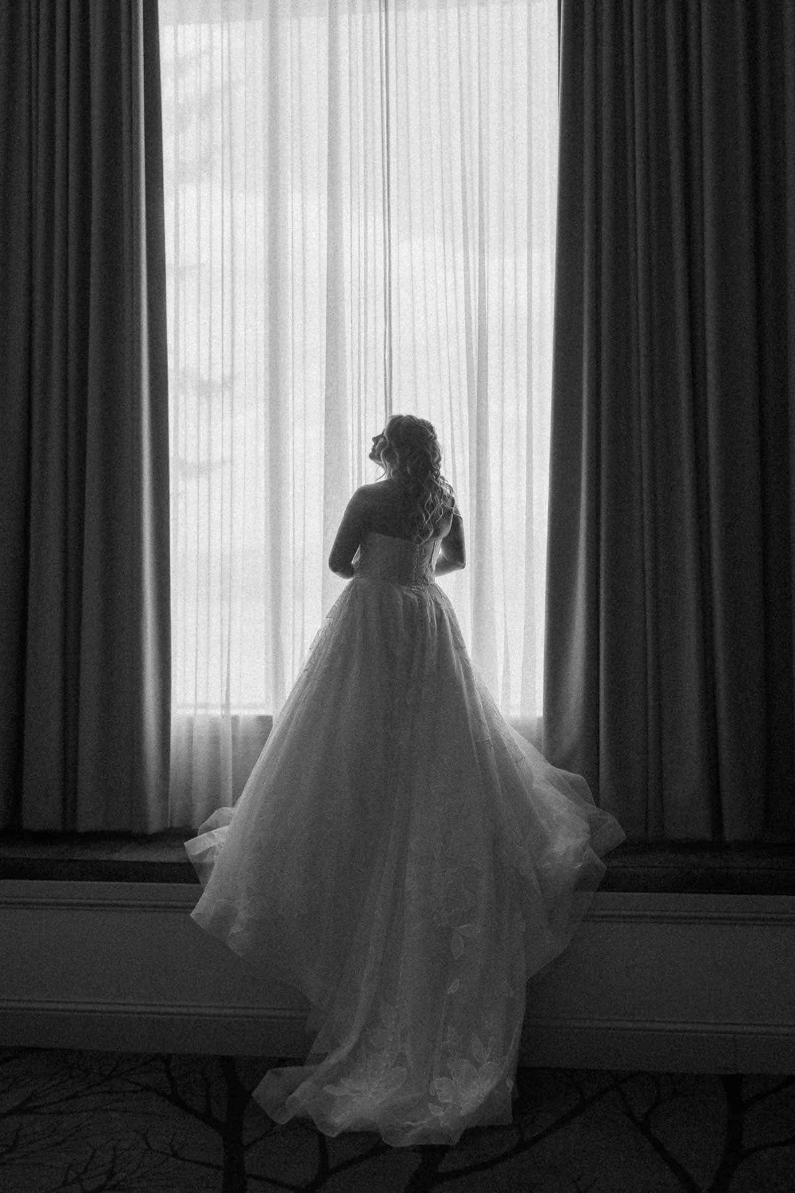 A woman in a flowing wedding dress stands with her back to the camera, gazing out a large window draped with sheer curtains, with soft light streaming in. The image is in black and white.
