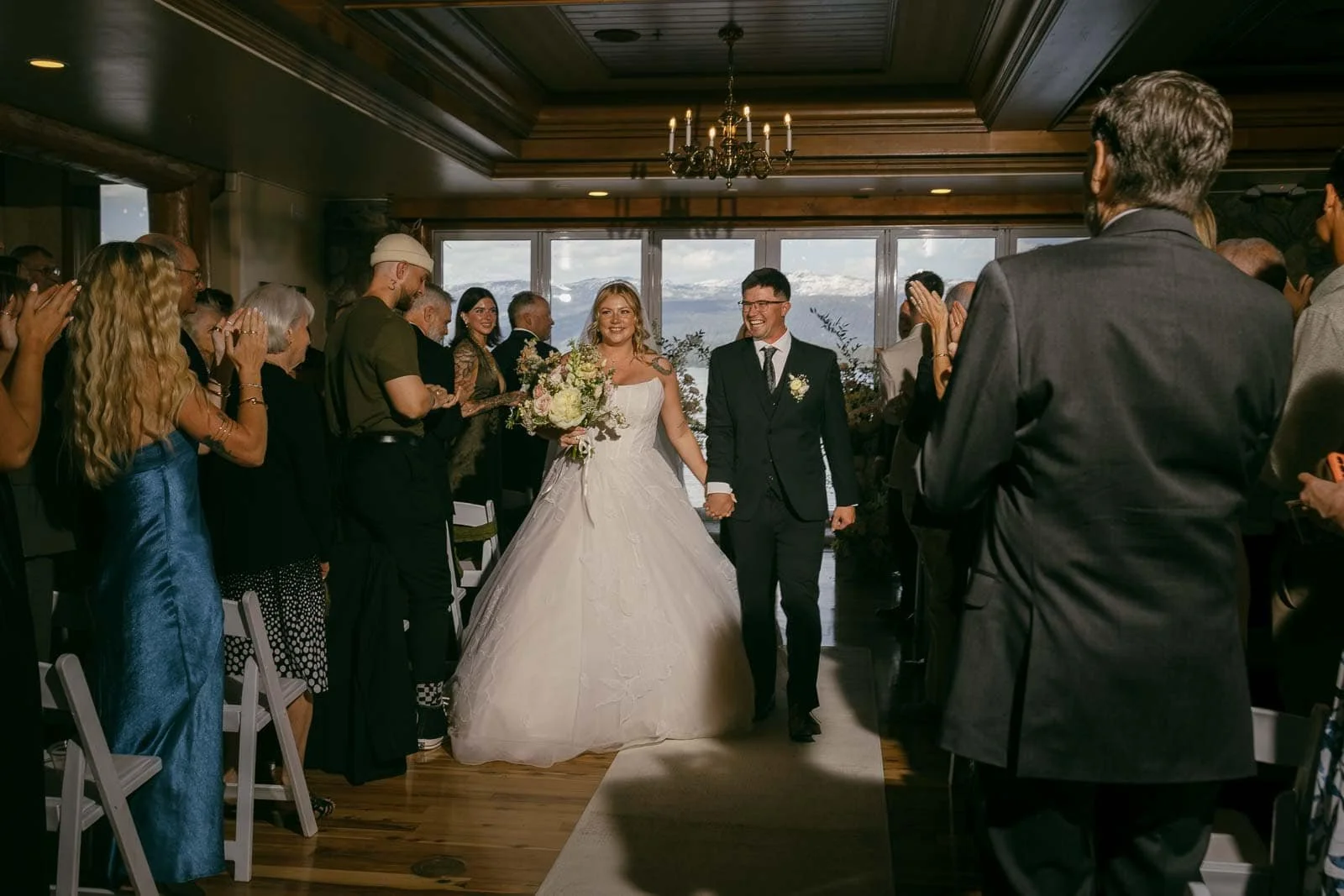 A bride in a white gown and a groom in a black suit walk down the aisle together, smiling, as guests on both sides stand and applaud in a warmly lit indoor venue.