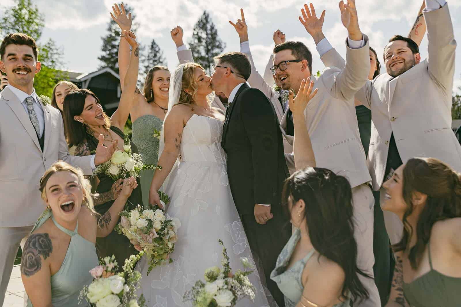 A bride and groom kiss as their joyful wedding party, dressed in pastels, surrounds them, cheering and raising their hands. Everyone is smiling and celebrating outdoors in sunlight.
