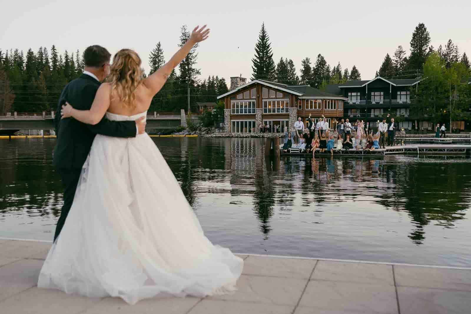 A bride and groom stand by a lakeside, arms around each other, waving to a group of people gathered on a dock across the water in front of Shore Lodge surrounded by trees.