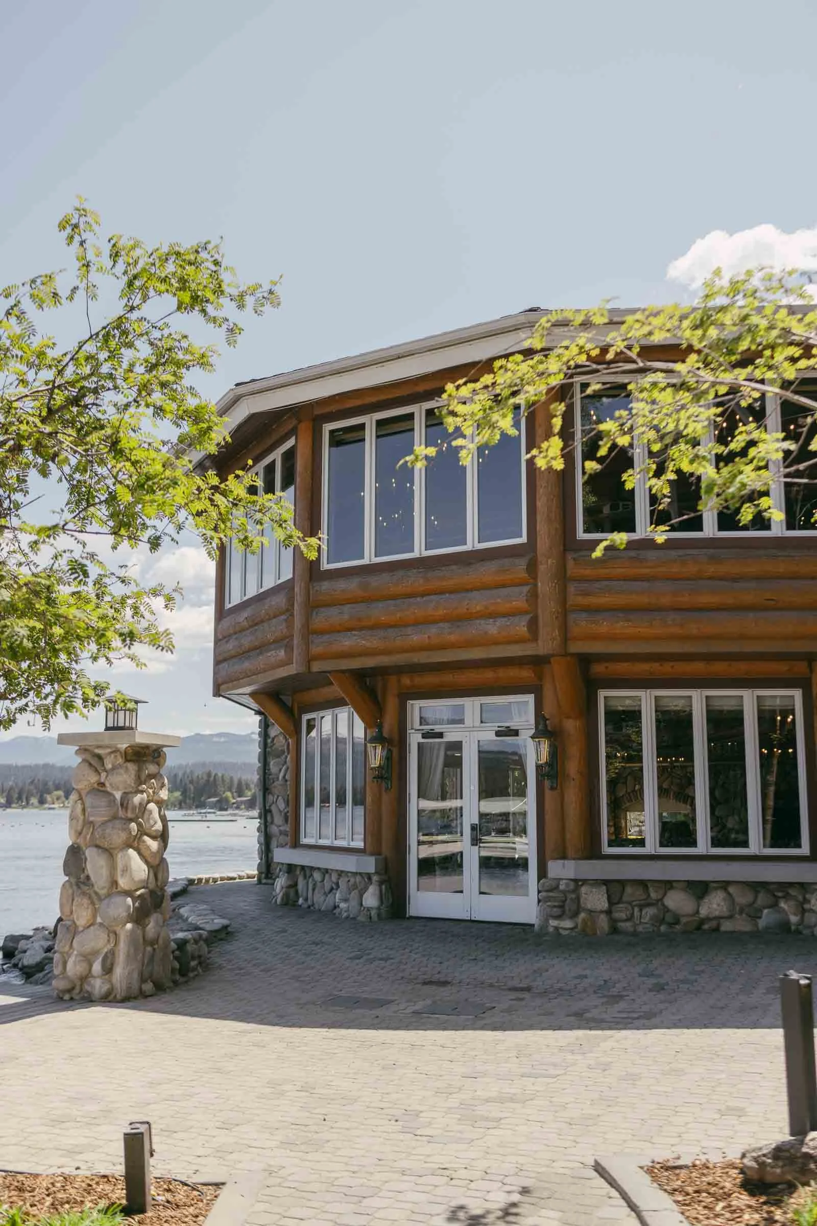  A round, two-story wooden building with large windows sits by a lake, surrounded by stone pillars and trees, under a clear sky with some clouds. The ceremony and reception space at Shore Lodge 
